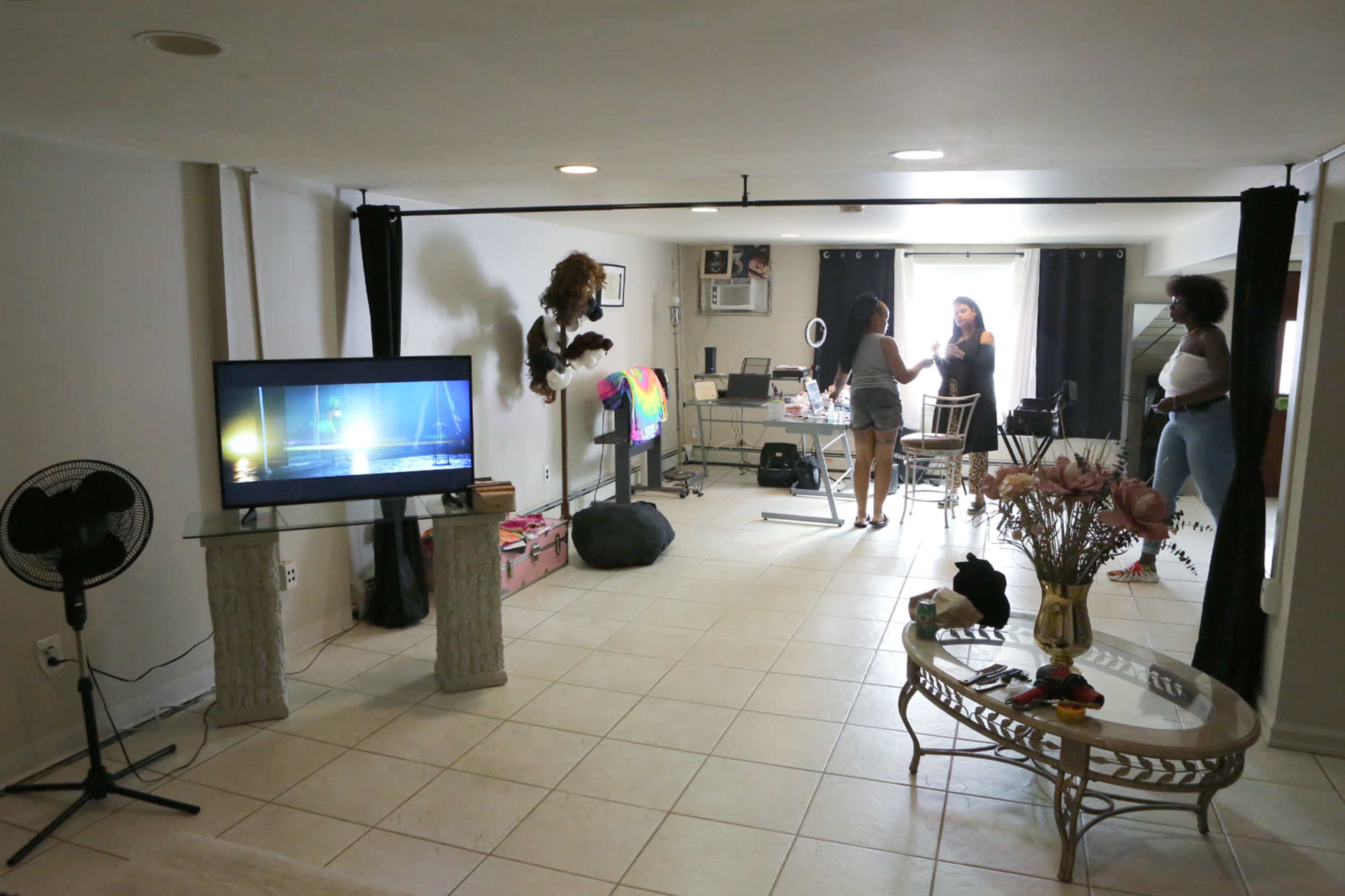 The image shows a spacious room featuring a television, a table with chairs, and two people standing near a makeup station, with a lion statue and decorative flowers in the foreground.