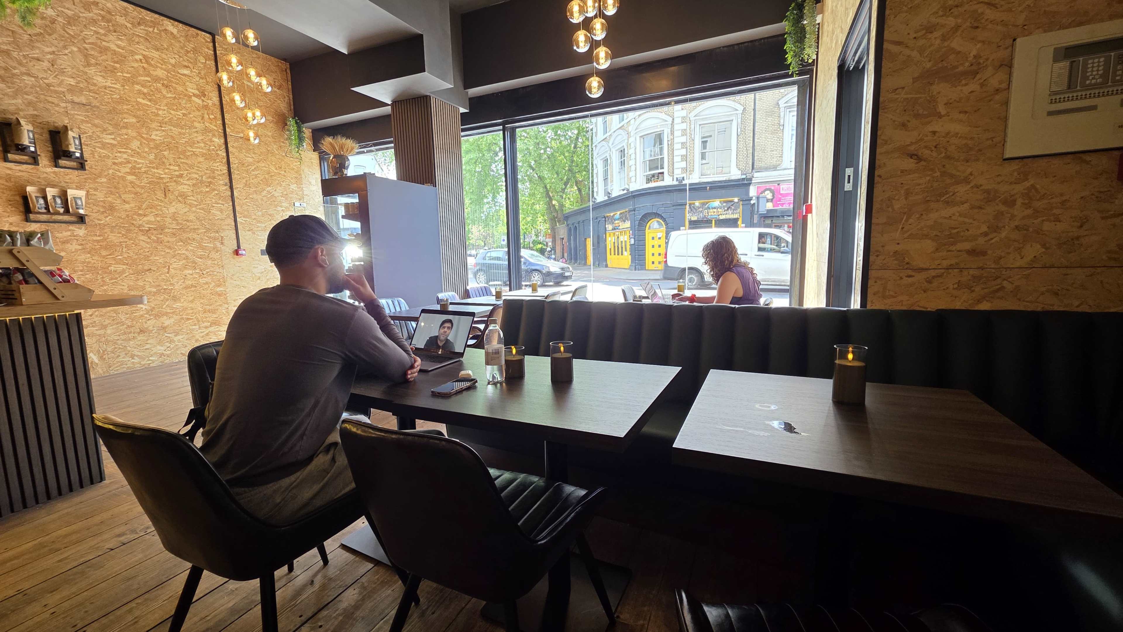 A person sits at a table in a café, looking at a device, while another person works in the background near a large window.