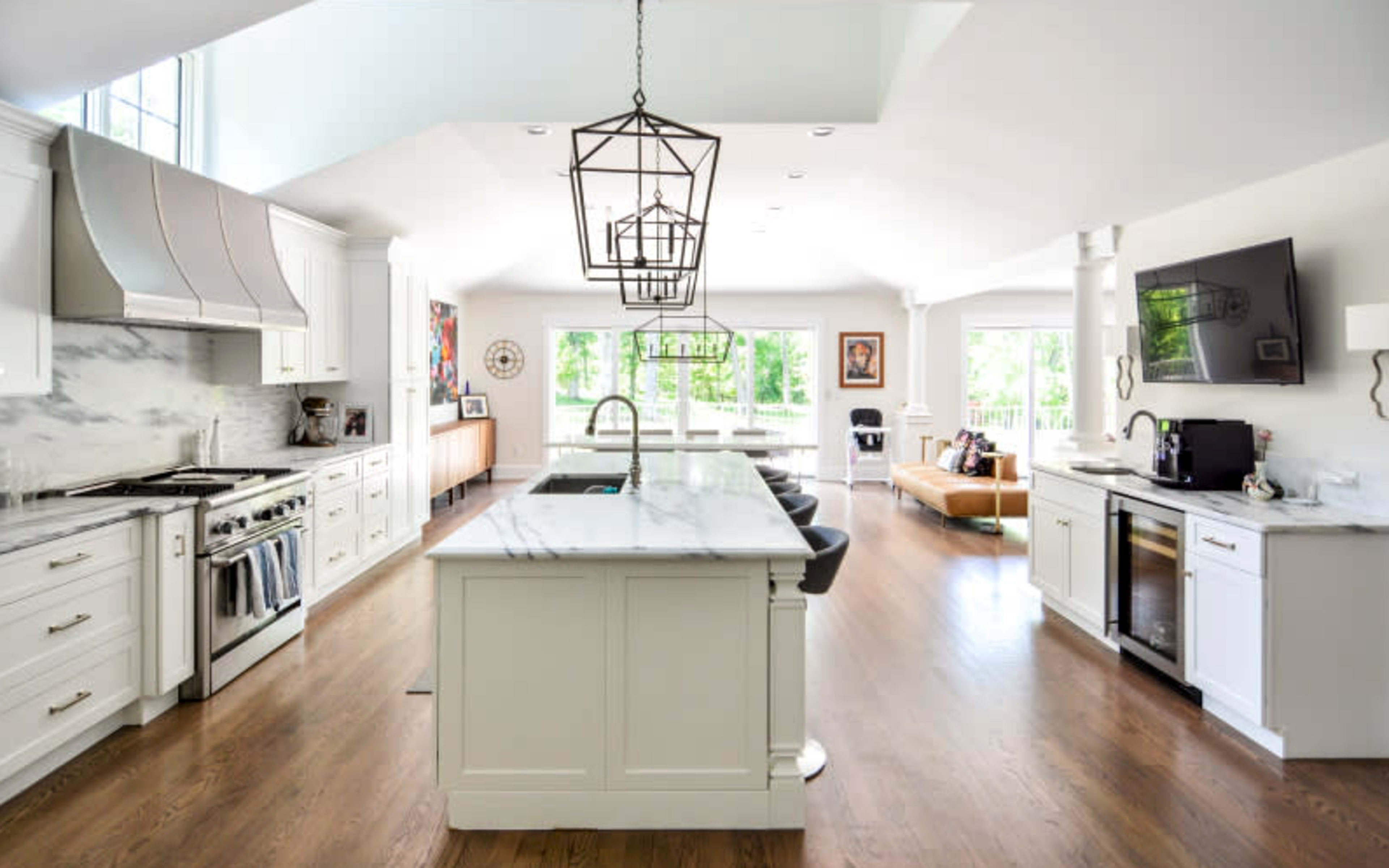 The image shows a modern kitchen with white cabinets, a large marble island, and an open layout that connects to a living area filled with natural light.