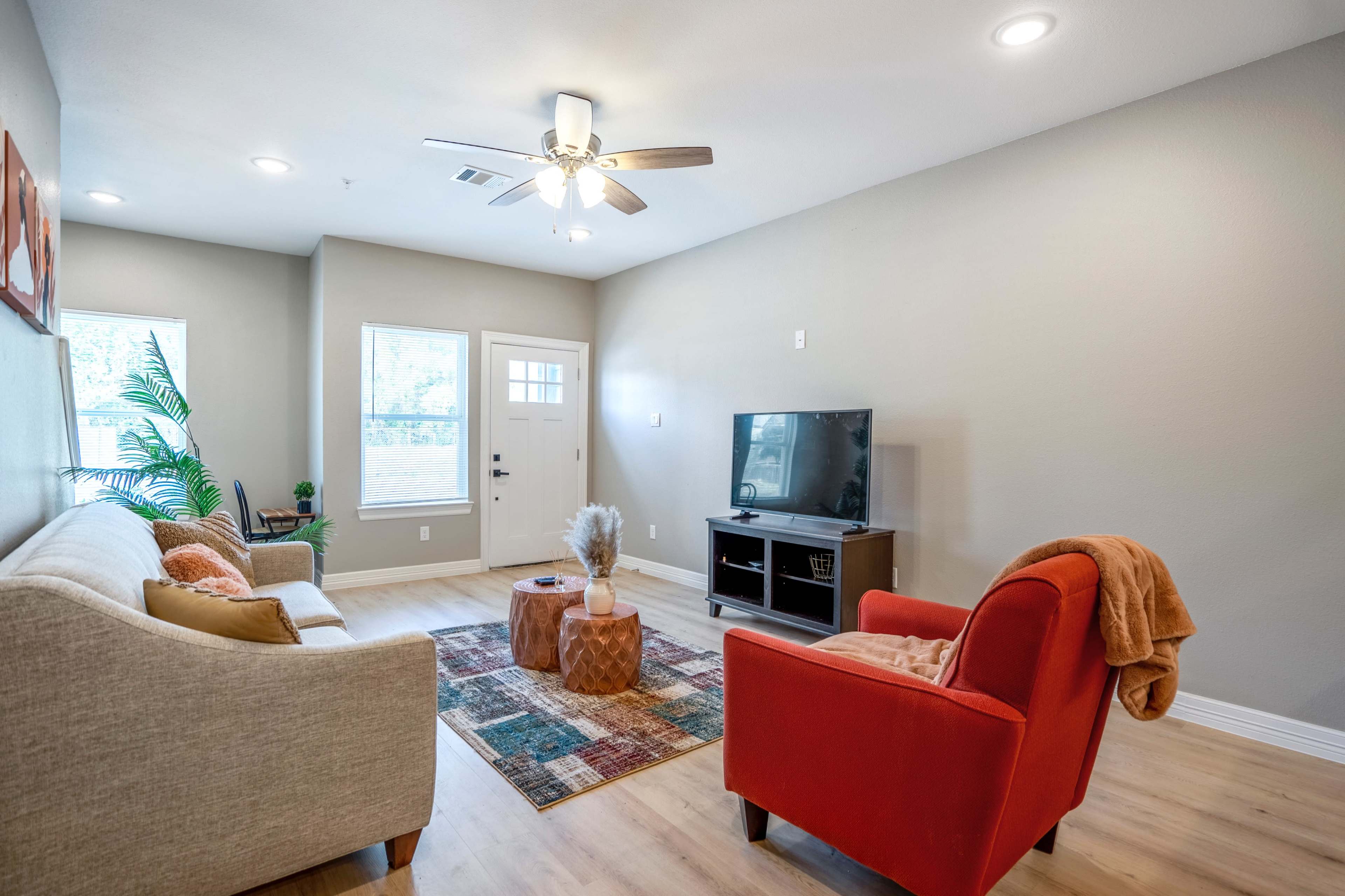 The living room features a beige sofa, a red armchair, a wooden TV stand, and a colorful area rug beneath a ceiling fan and natural light from the windows.