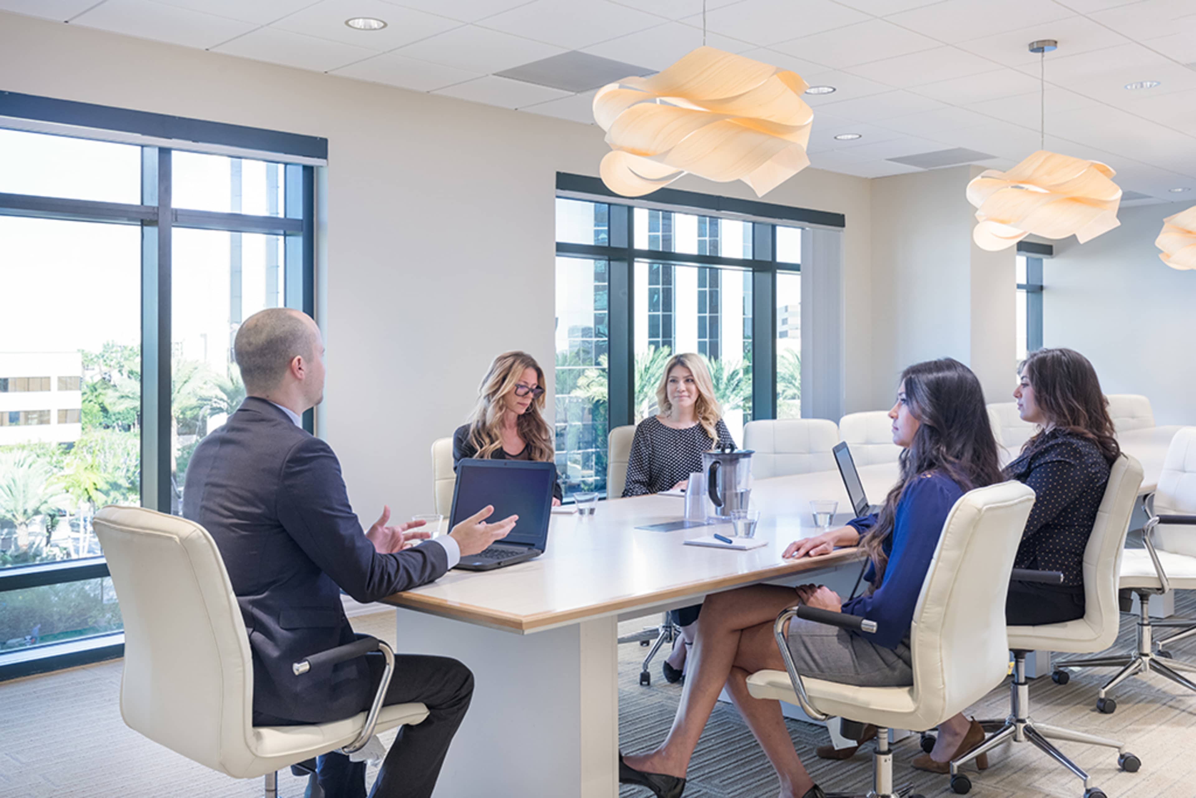 A group of five professionals engages in a discussion around a modern conference table in a well-lit office with large windows.