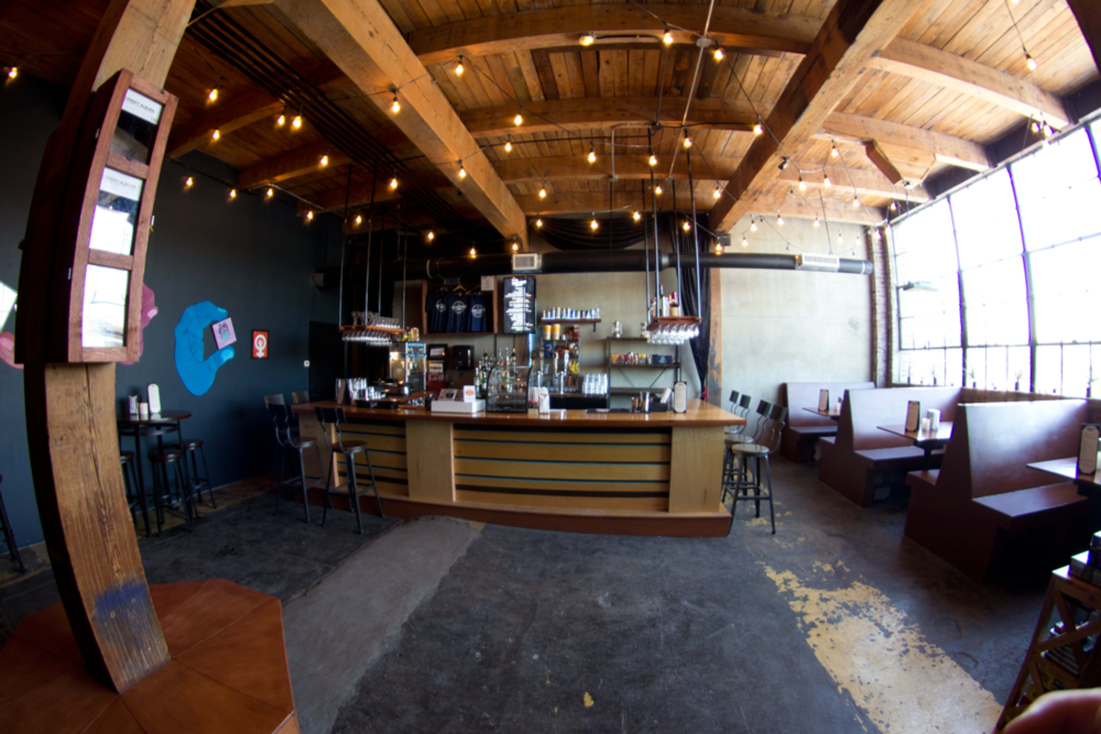 A spacious bar area with wooden beams, a long counter, and seating along the walls, illuminated by string lights.
