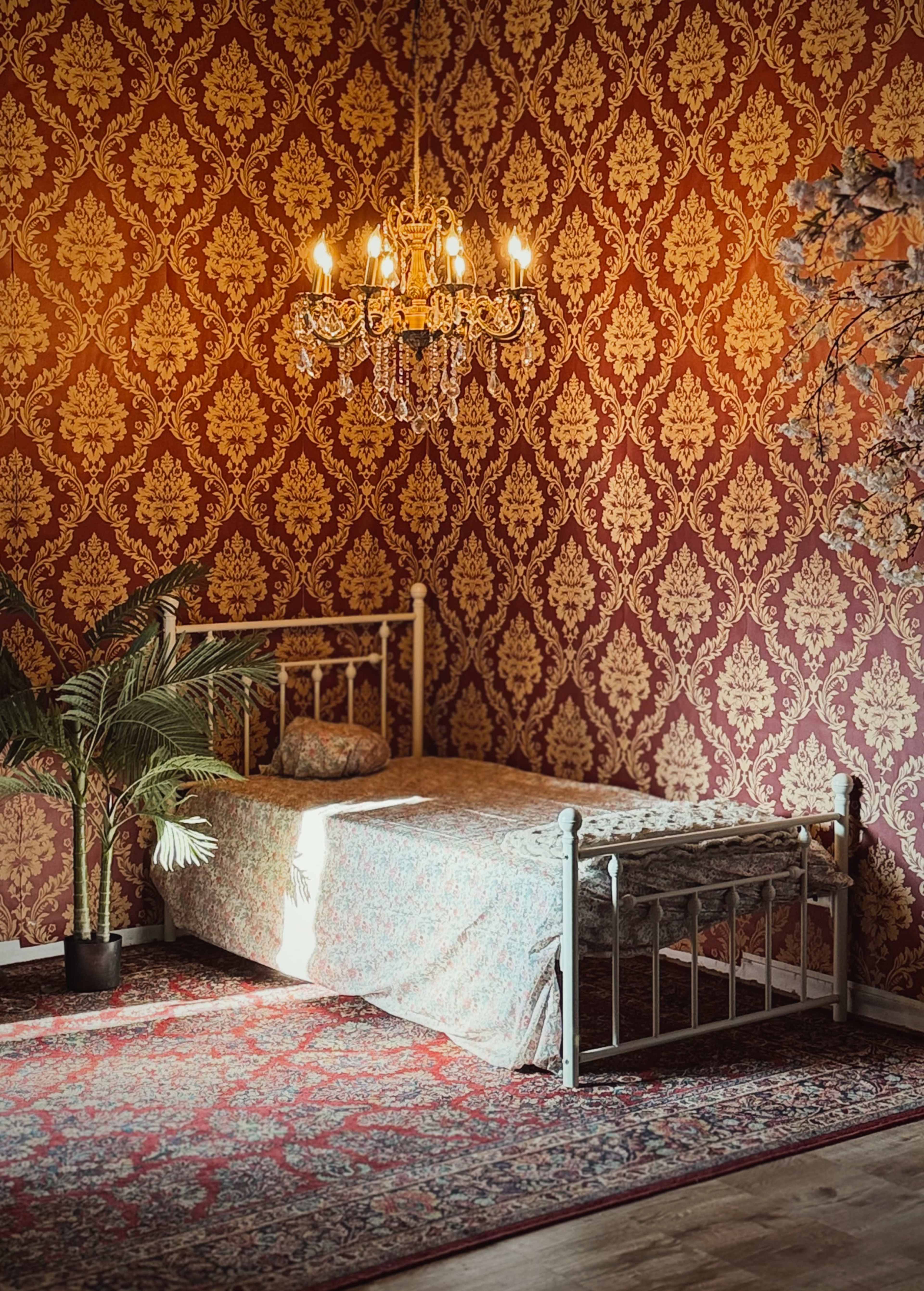 The image depicts a bedroom with a single bed, a patterned rug, a palm plant in a pot, and a chandelier hanging from the ceiling, all set against a richly patterned wall.
