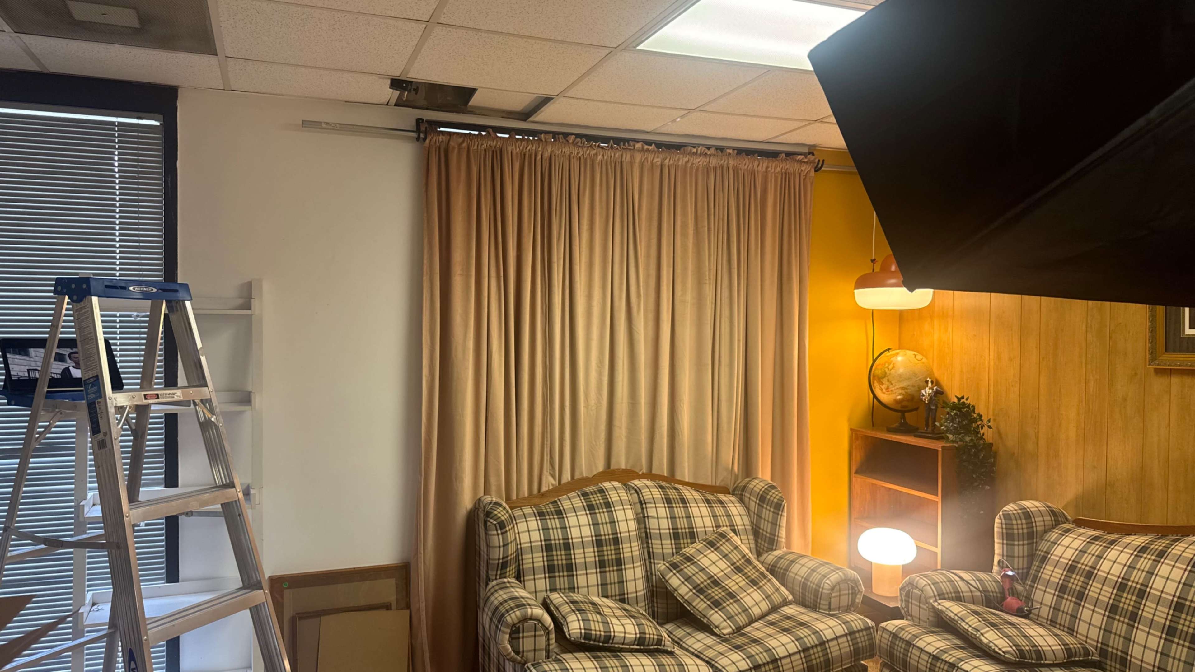 The image shows a cozy room with two patterned sofas, a floor lamp, a globe on a shelf, and a step ladder beside a window with curtains.