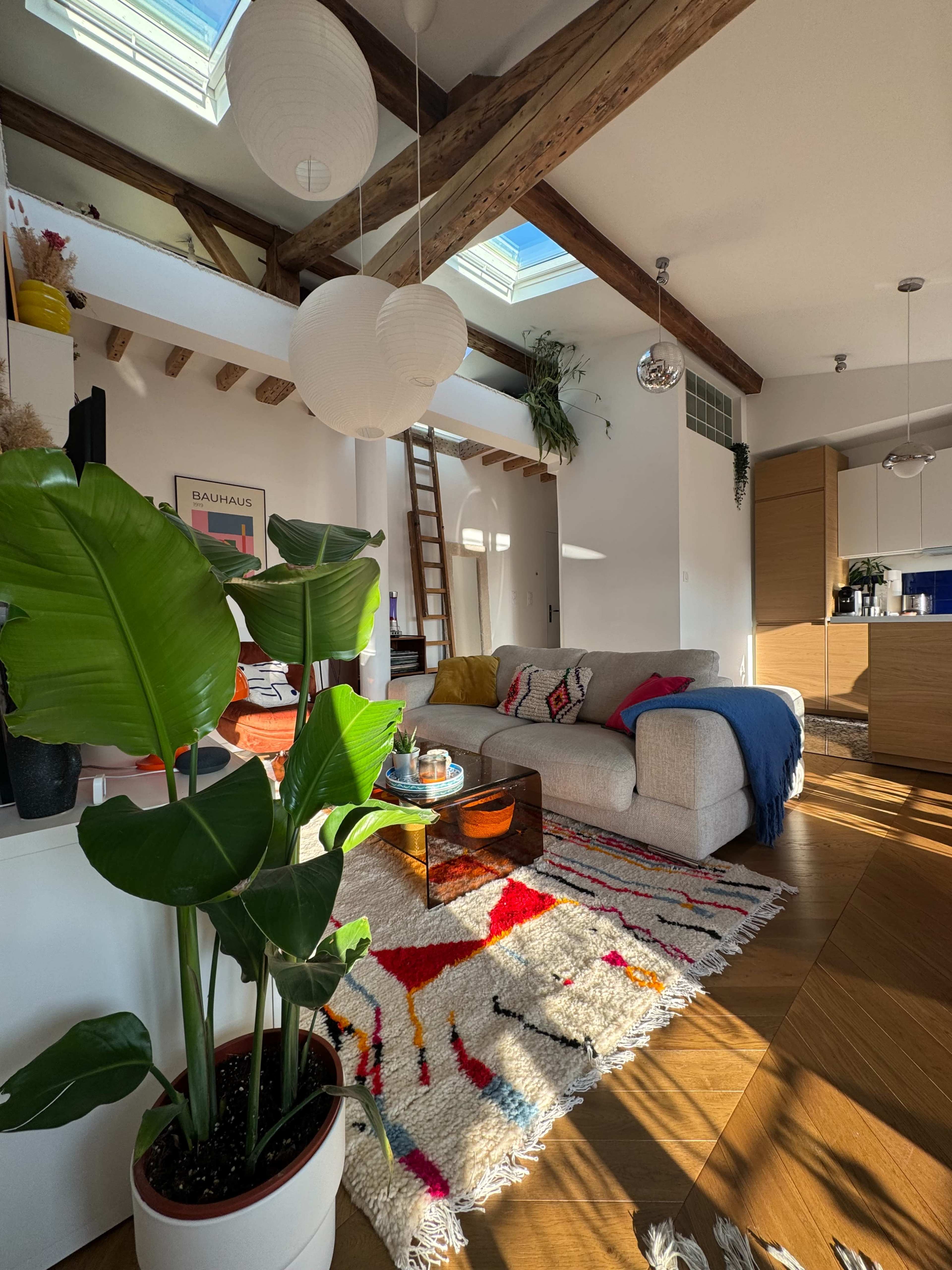 The image shows a cozy living room with a large sectional sofa, a colorful rug, and a plant, featuring wooden beams and skylights above.