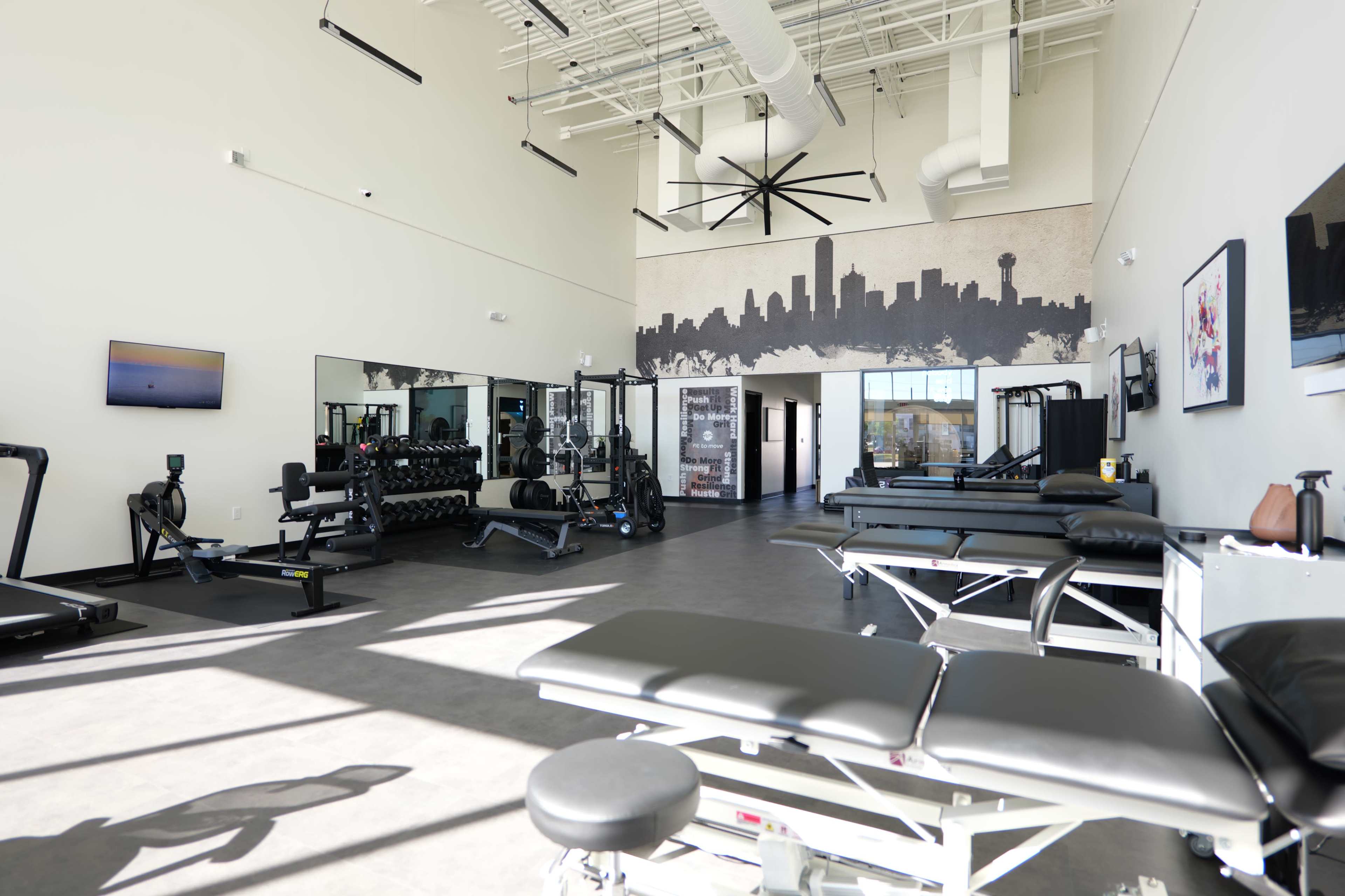 A spacious gym features various exercise equipment, treatment tables, and a skyline mural on the wall.