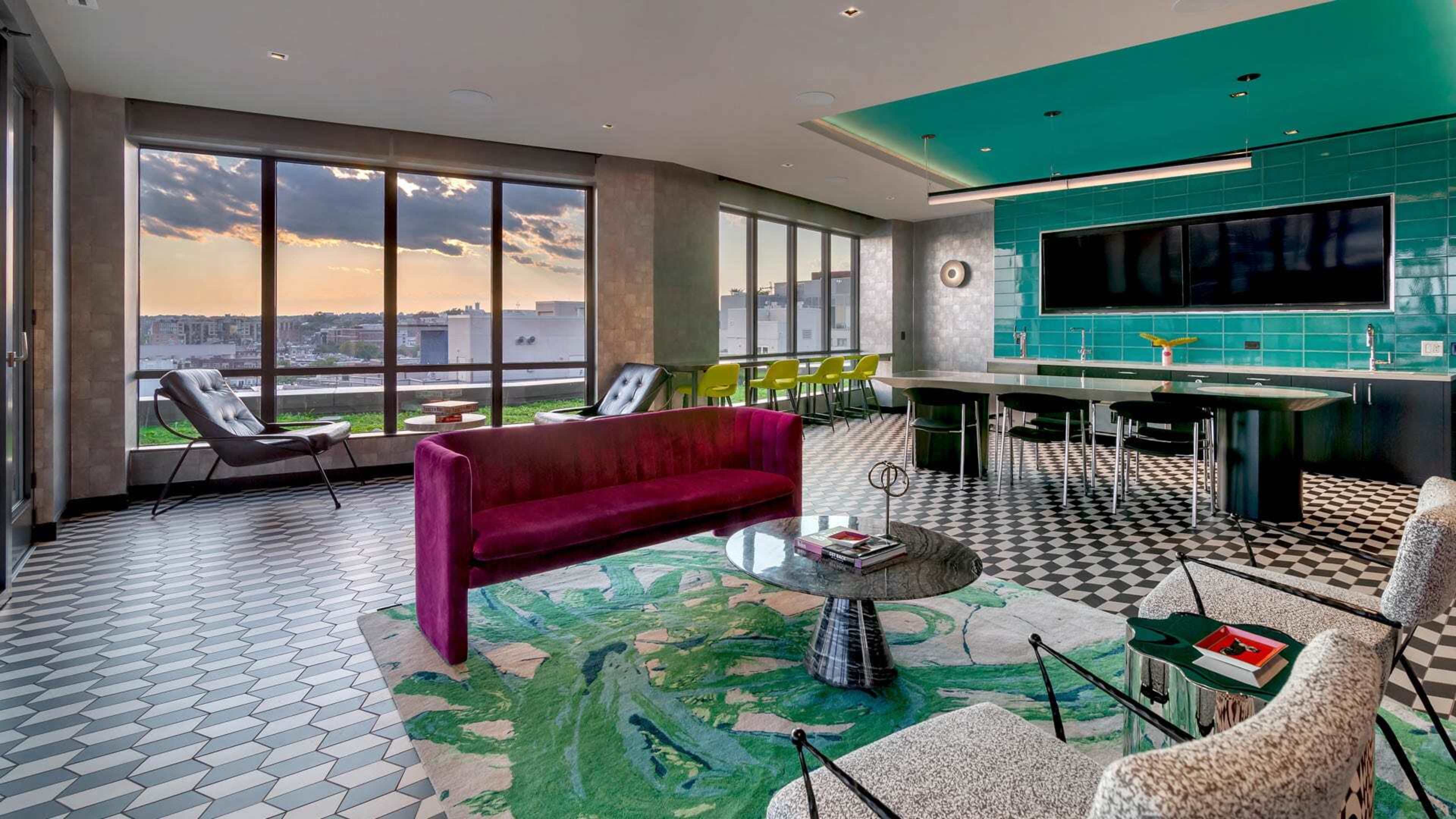 The image shows a modern, stylish interior with large windows, featuring a seating area with a vibrant sofa, patterned tiles, and a green-accented wall.