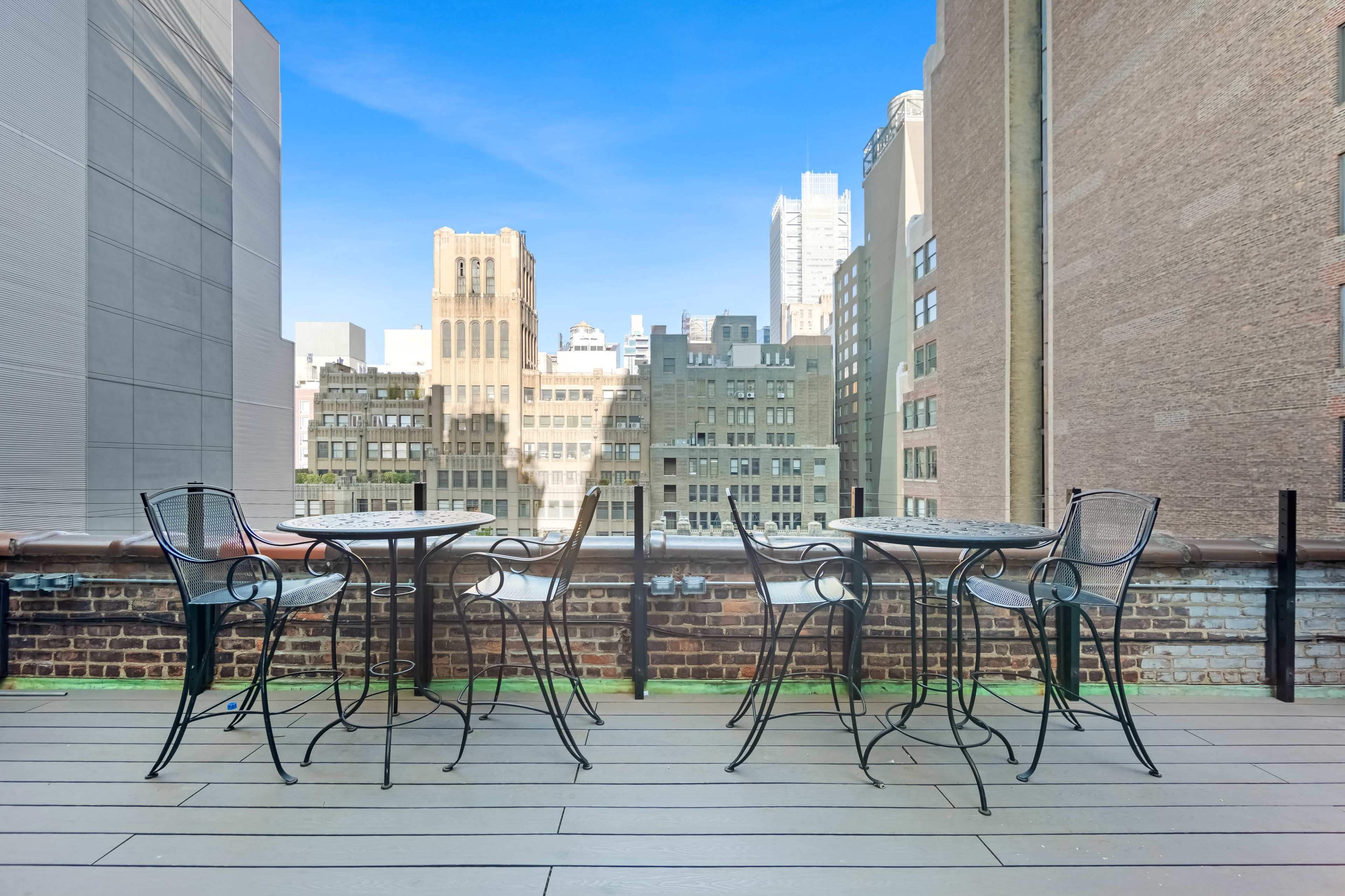 A rooftop terrace features several black metal bistro tables and chairs overlooking a city skyline with tall buildings.