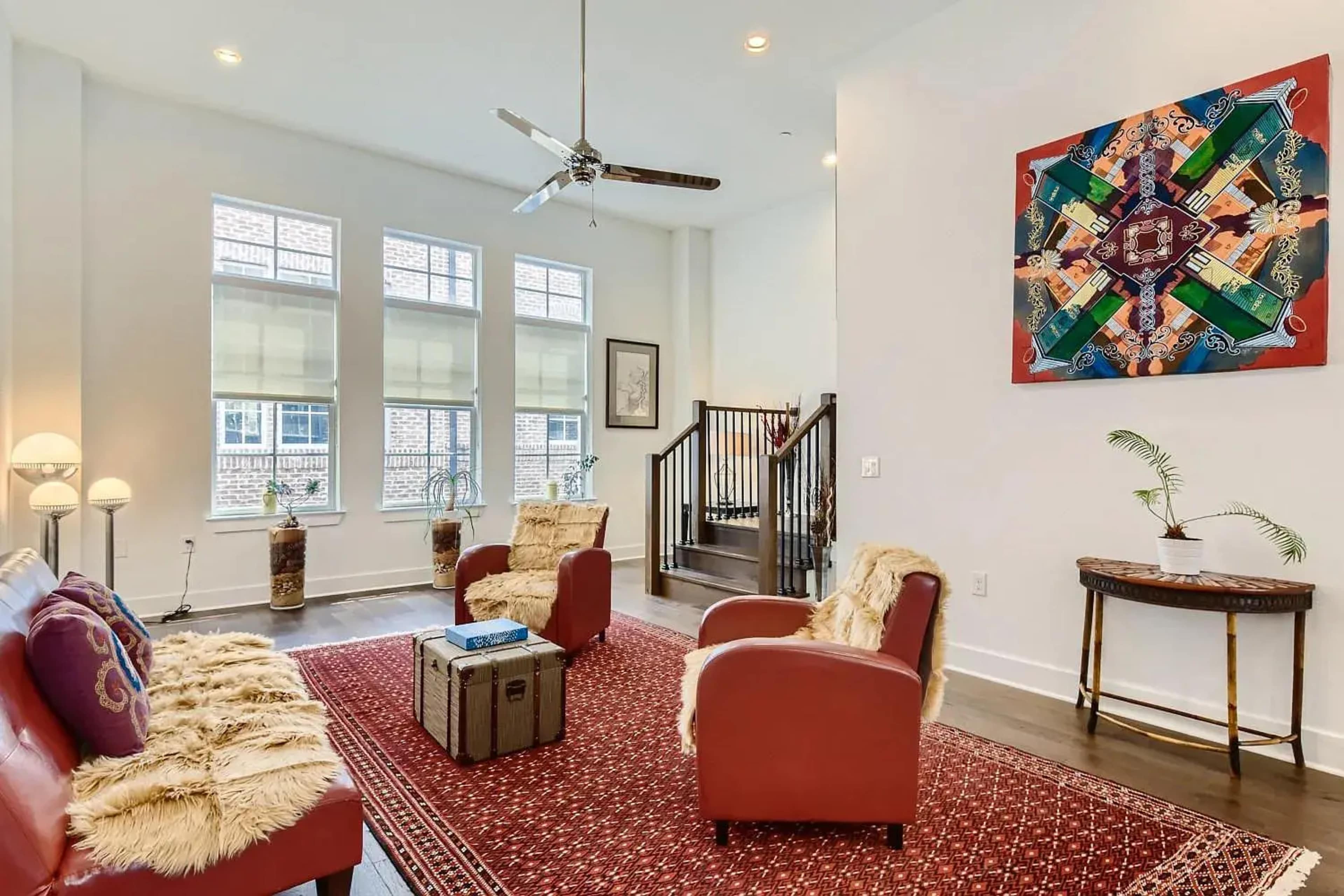 The image shows a bright living room with two red armchairs, a red sofa with decorative pillows, a large wall-mounted artwork, and a patterned rug on a hardwood floor.