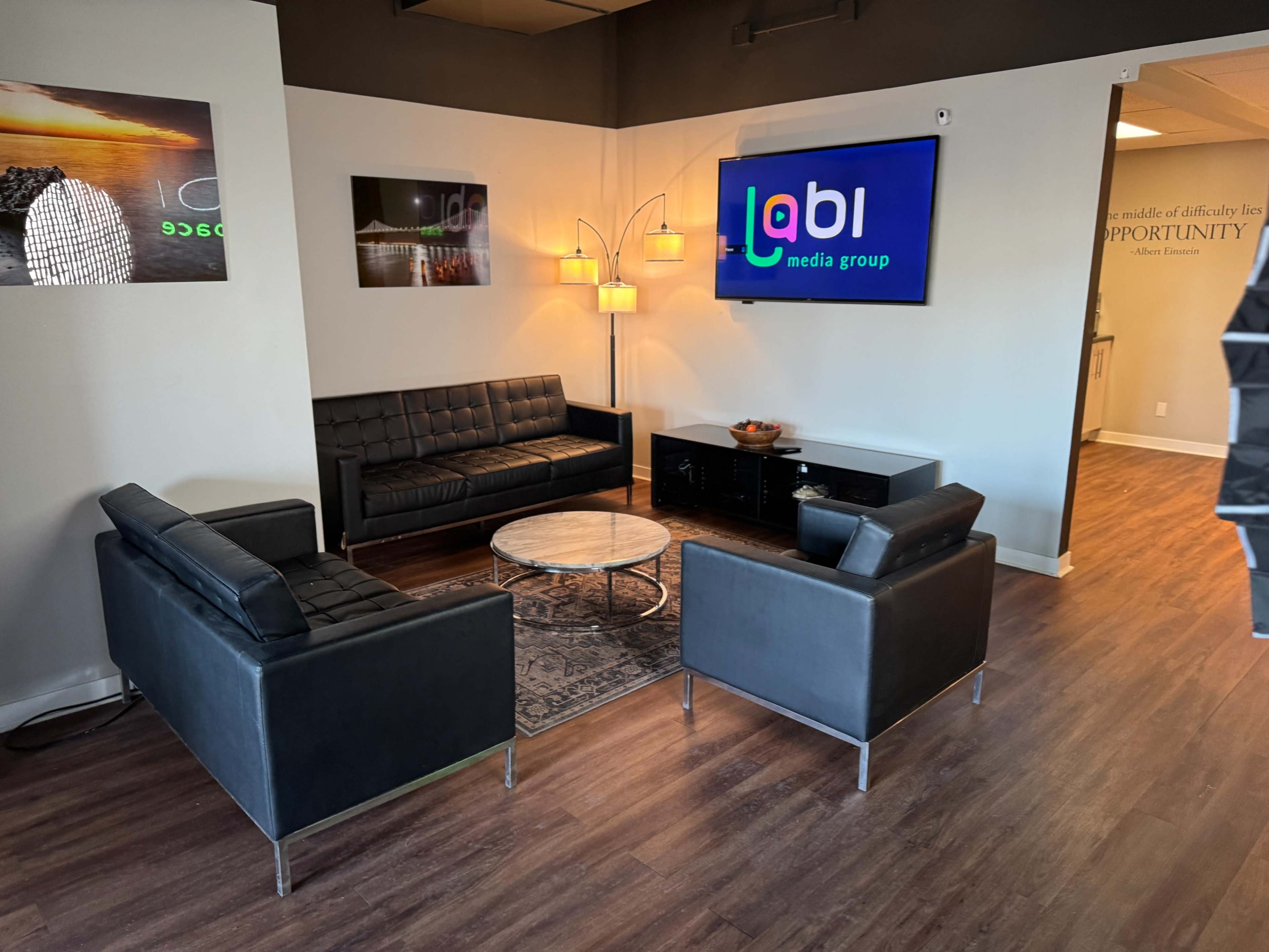 The image shows a modern office lounge area with black couches, a glass coffee table, and a wall-mounted television displaying the logo for a media group.