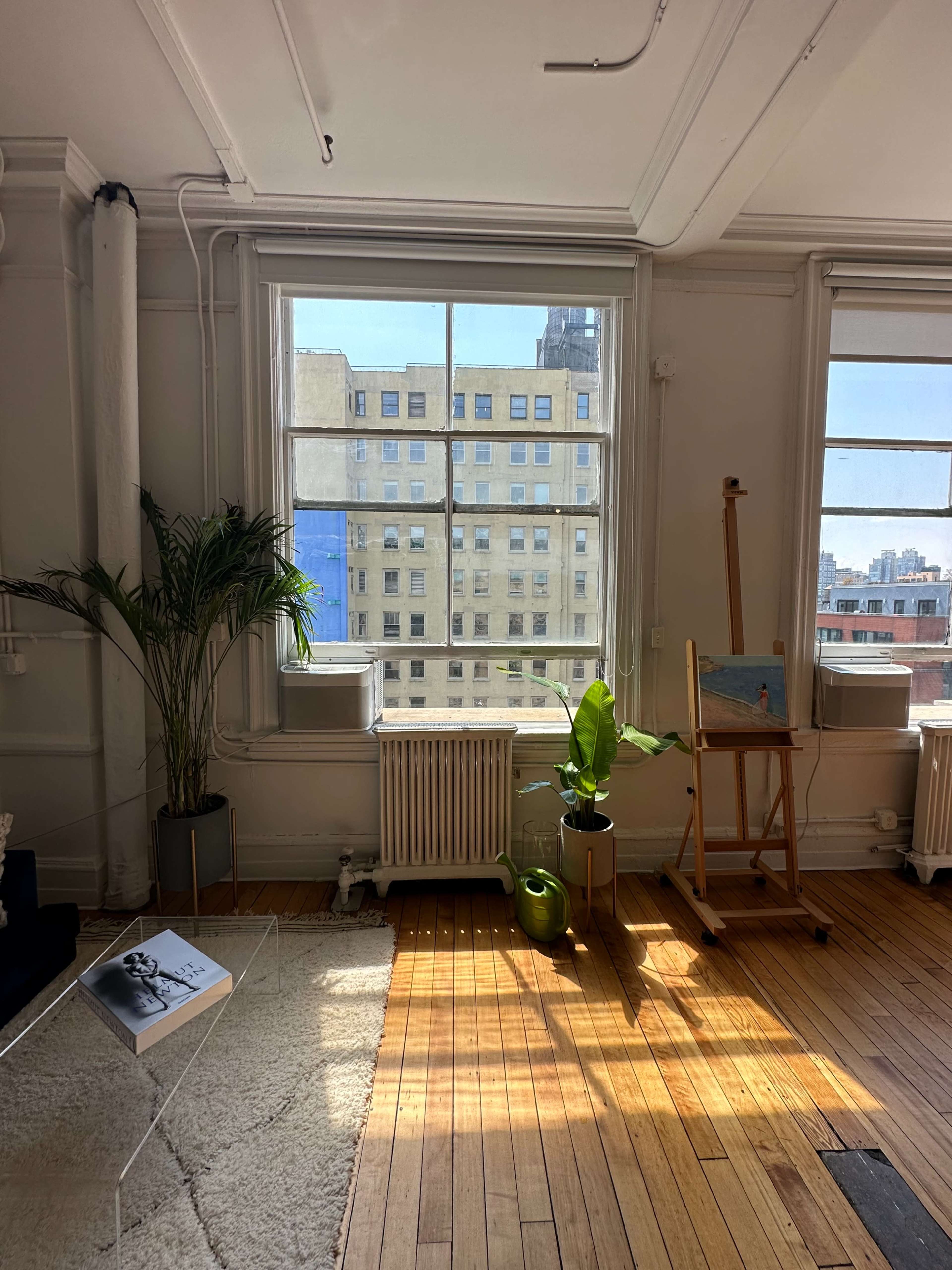 The image shows a bright room with large windows, a plant, an easel, and a small coffee table on a wooden floor.