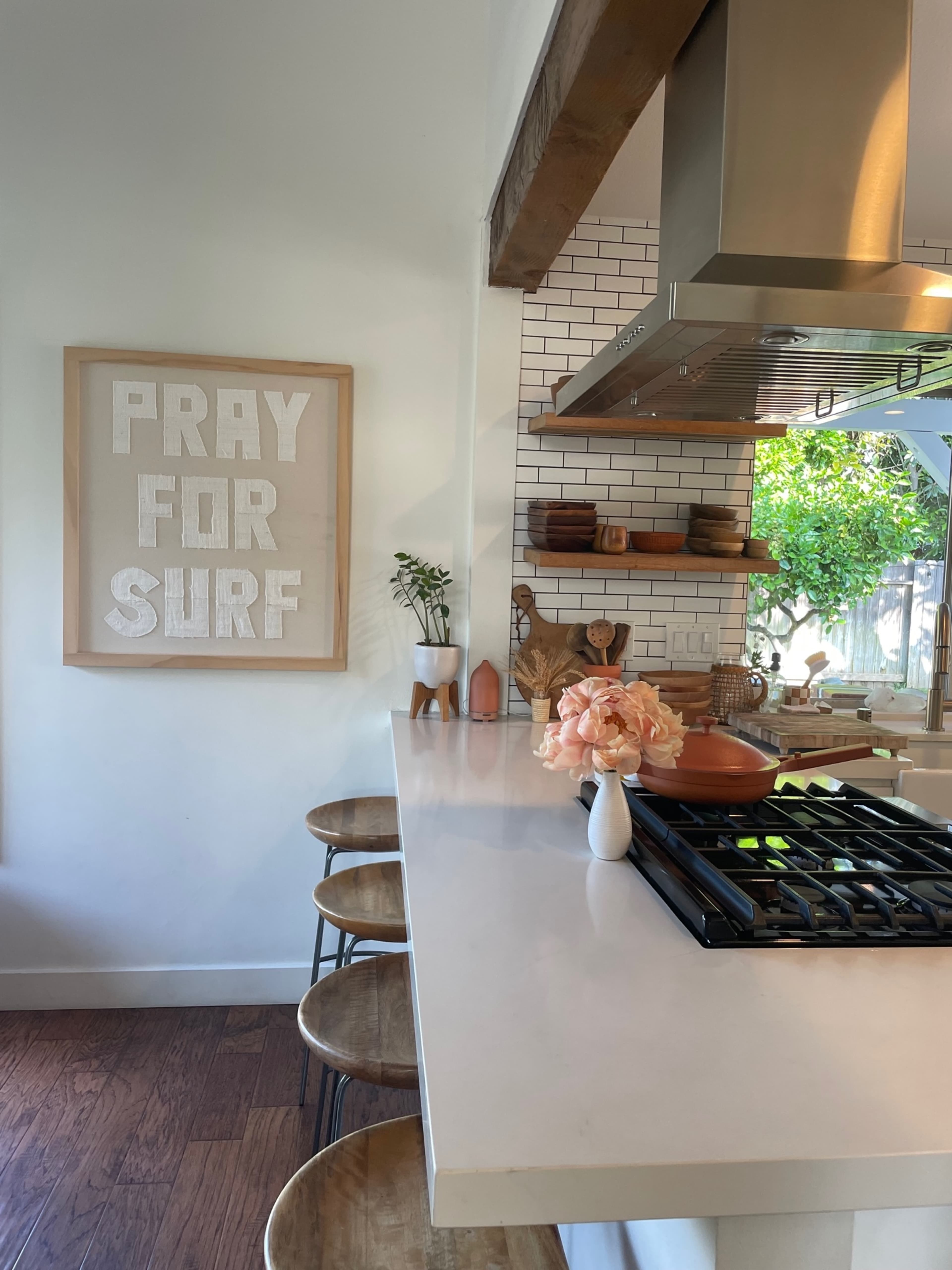 A modern kitchen features a white countertop with bar stools, a wall art piece that reads "PRAY FOR SURF," and a view of greenery through a window.