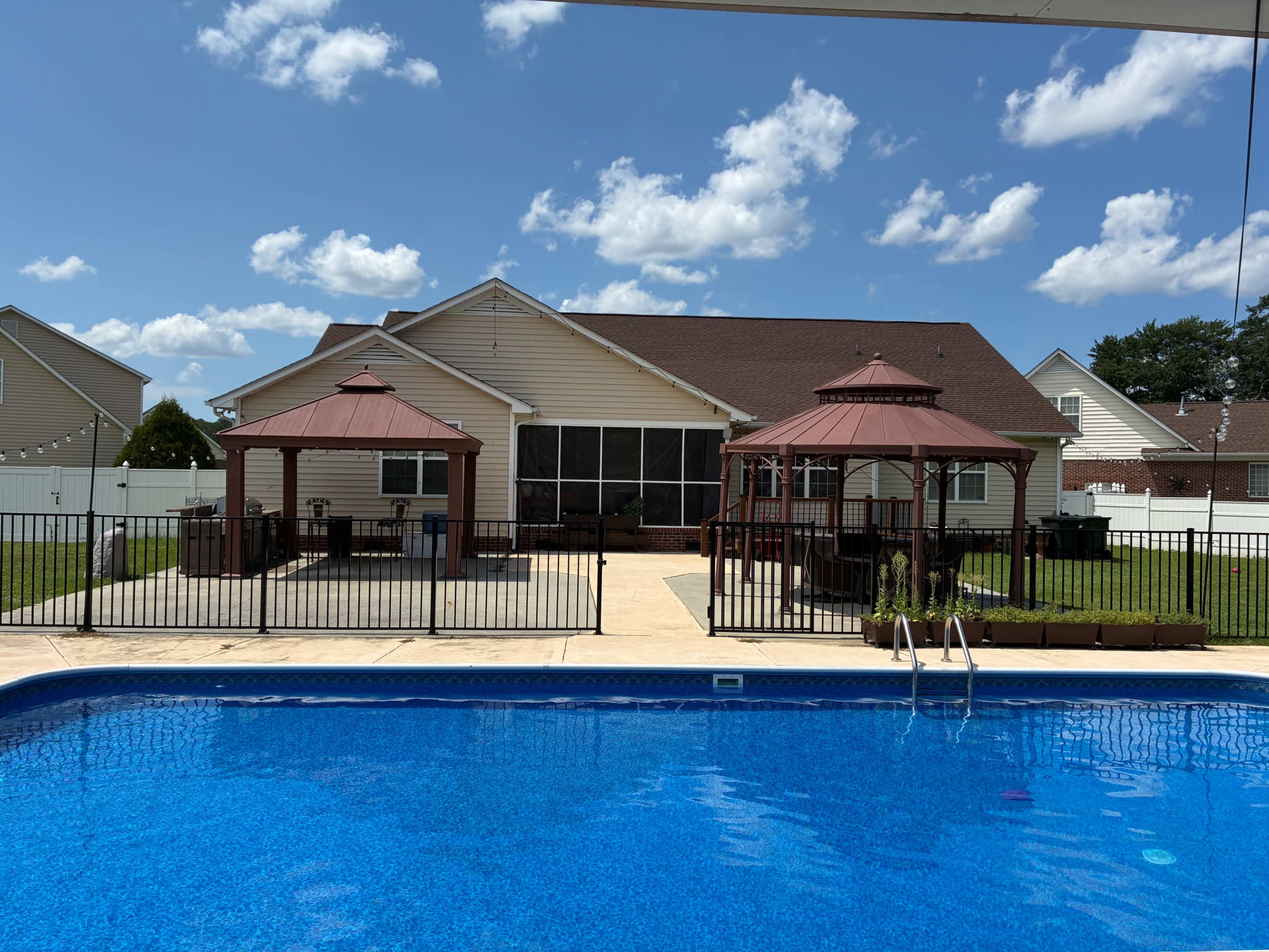 The image shows a backyard with a blue swimming pool, two gazebo structures, and a house in the background under a partly cloudy sky.