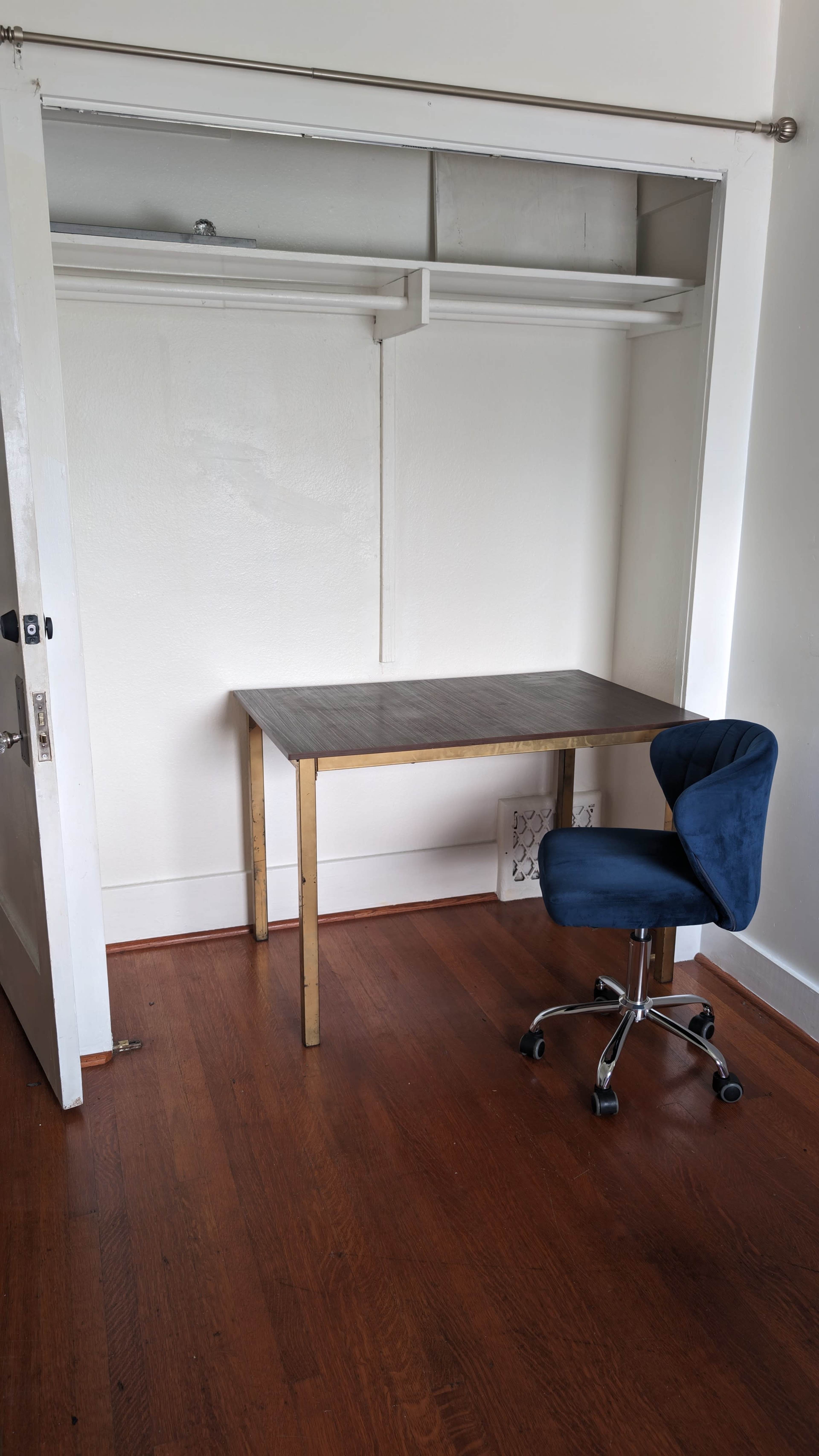 A simple room features a wooden desk and a blue swivel chair positioned next to an open closet.