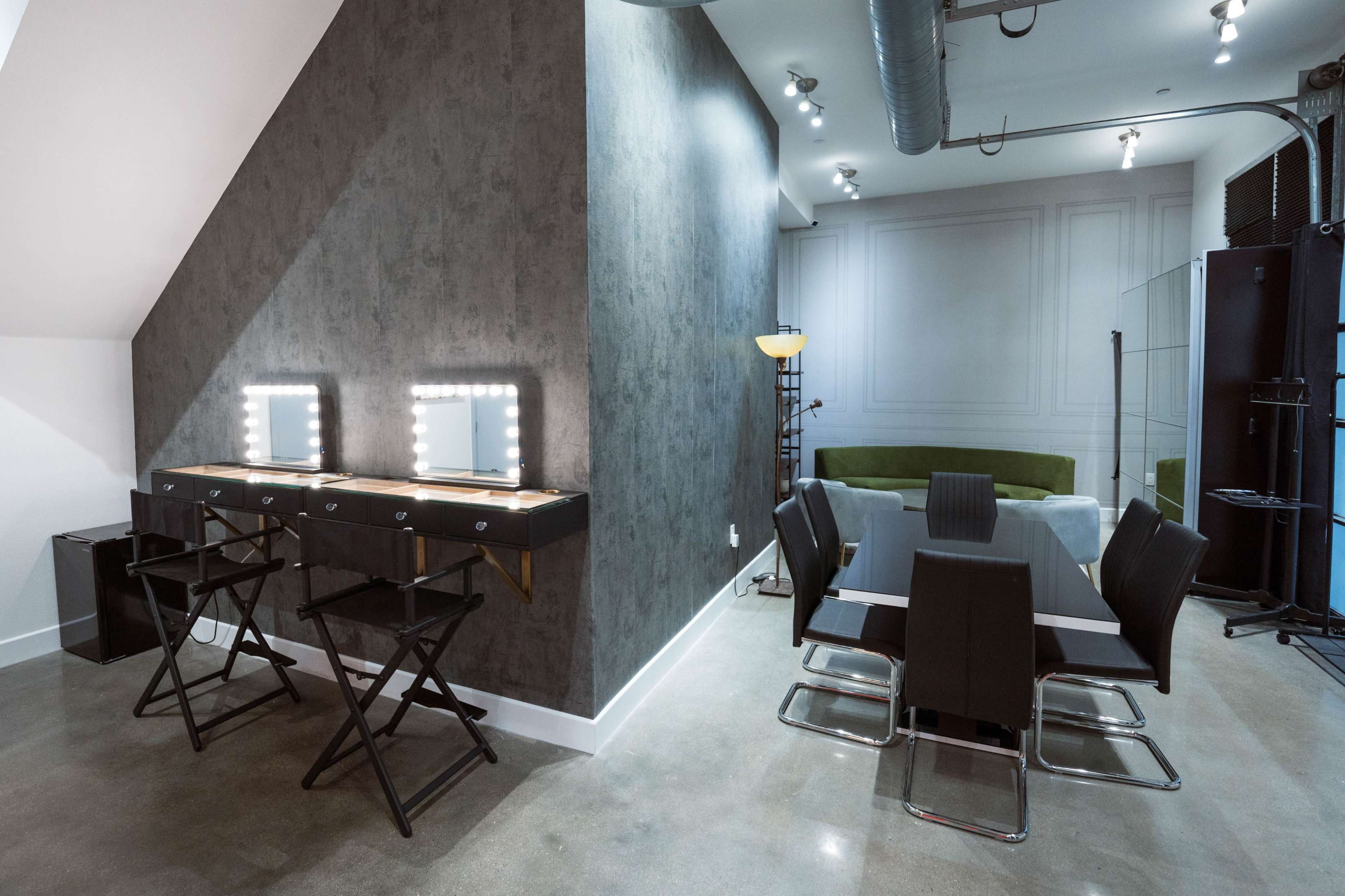 A modern room with two lighted vanity mirrors on a table, black folding chairs, and a seating area featuring a green couch and dining chairs.