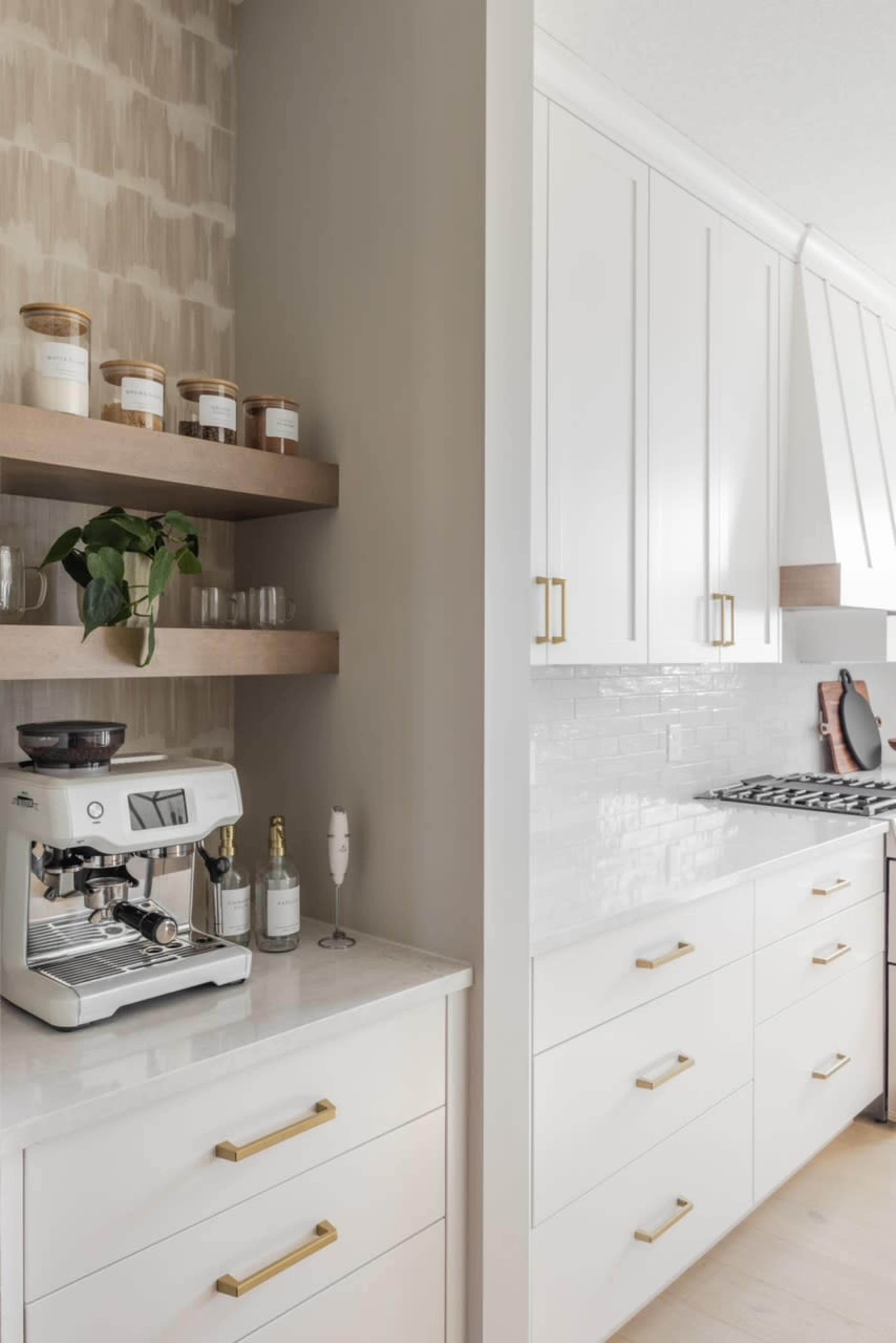 The image shows a modern kitchen with white cabinetry, a coffee machine on a countertop, and glass jars displayed on wooden shelves.