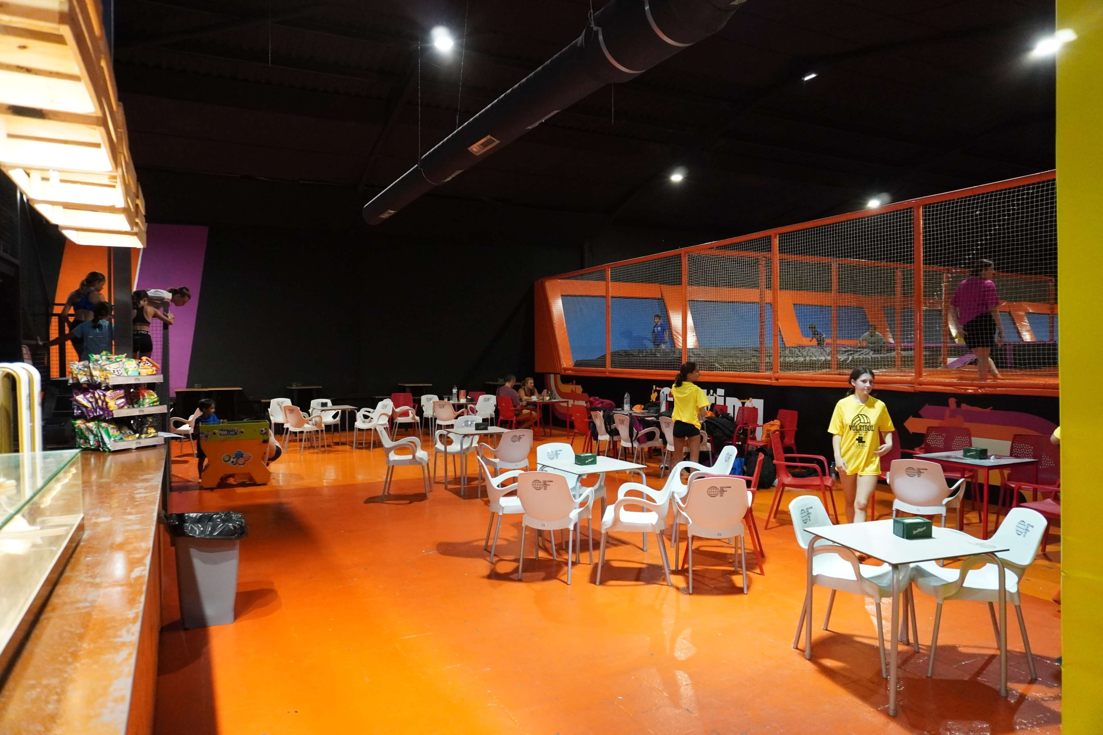 An indoor trampoline park with an adjacent seating area featuring brightly colored chairs and tables, while people can be seen jumping on trampolines behind a safety net.