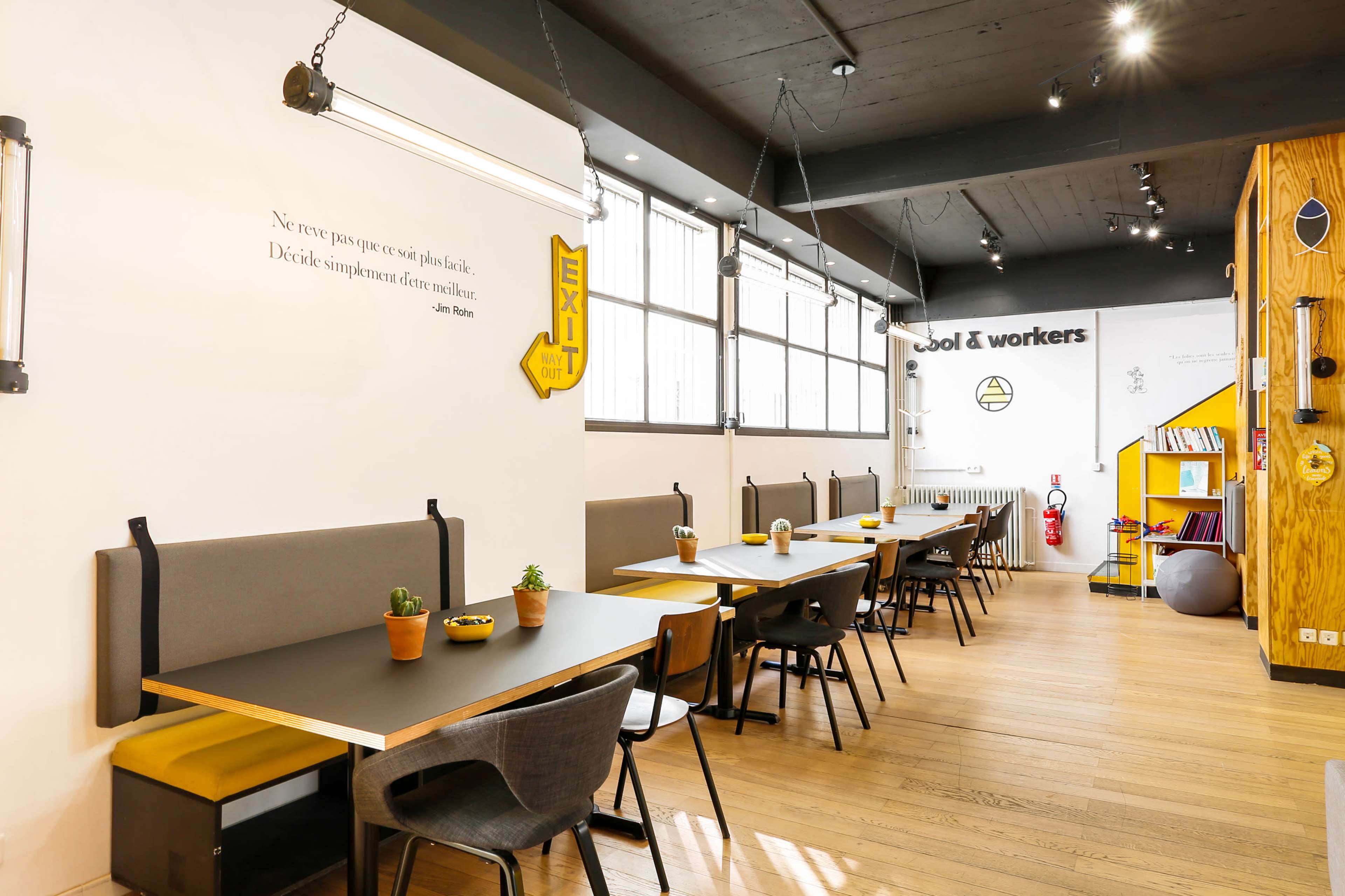 The image shows a modern workspace with long tables, potted plants, and a large exit sign on the wall.