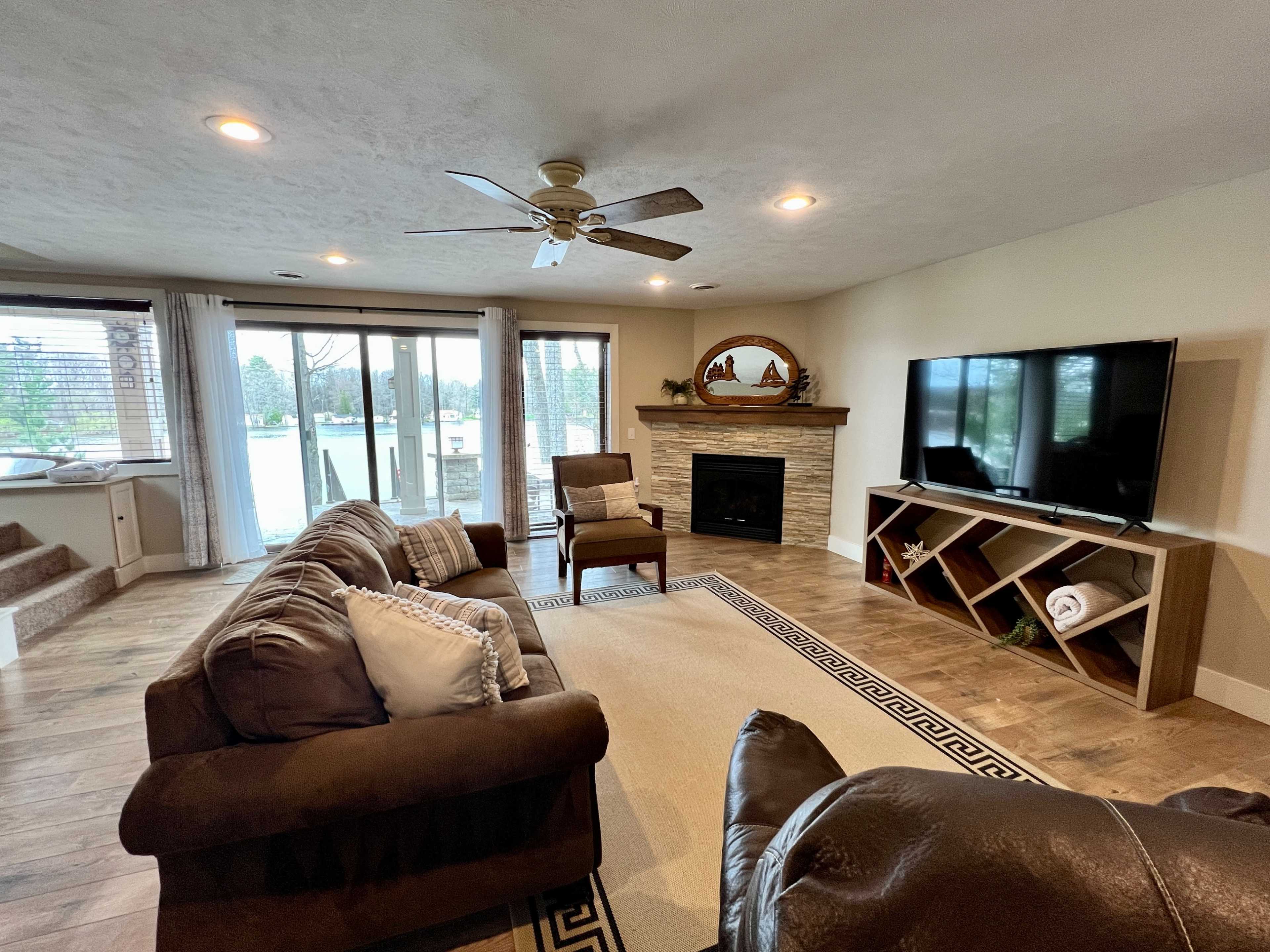 The image shows a cozy living room with a stone fireplace, large windows, a television, and comfortable seating around a neutral-colored area rug.