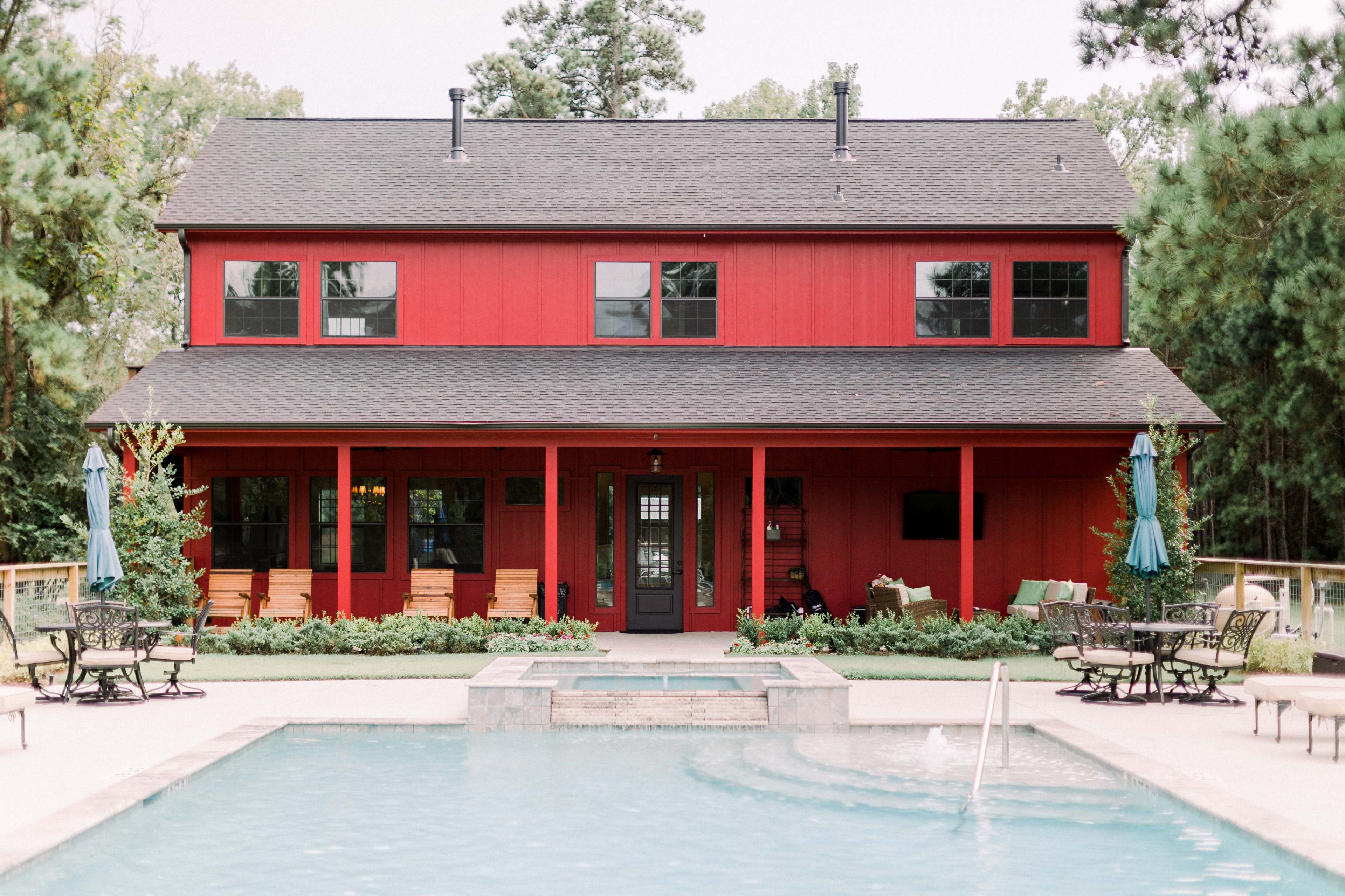 A two-story red house with a covered porch is situated next to a swimming pool surrounded by patio furniture and umbrellas.