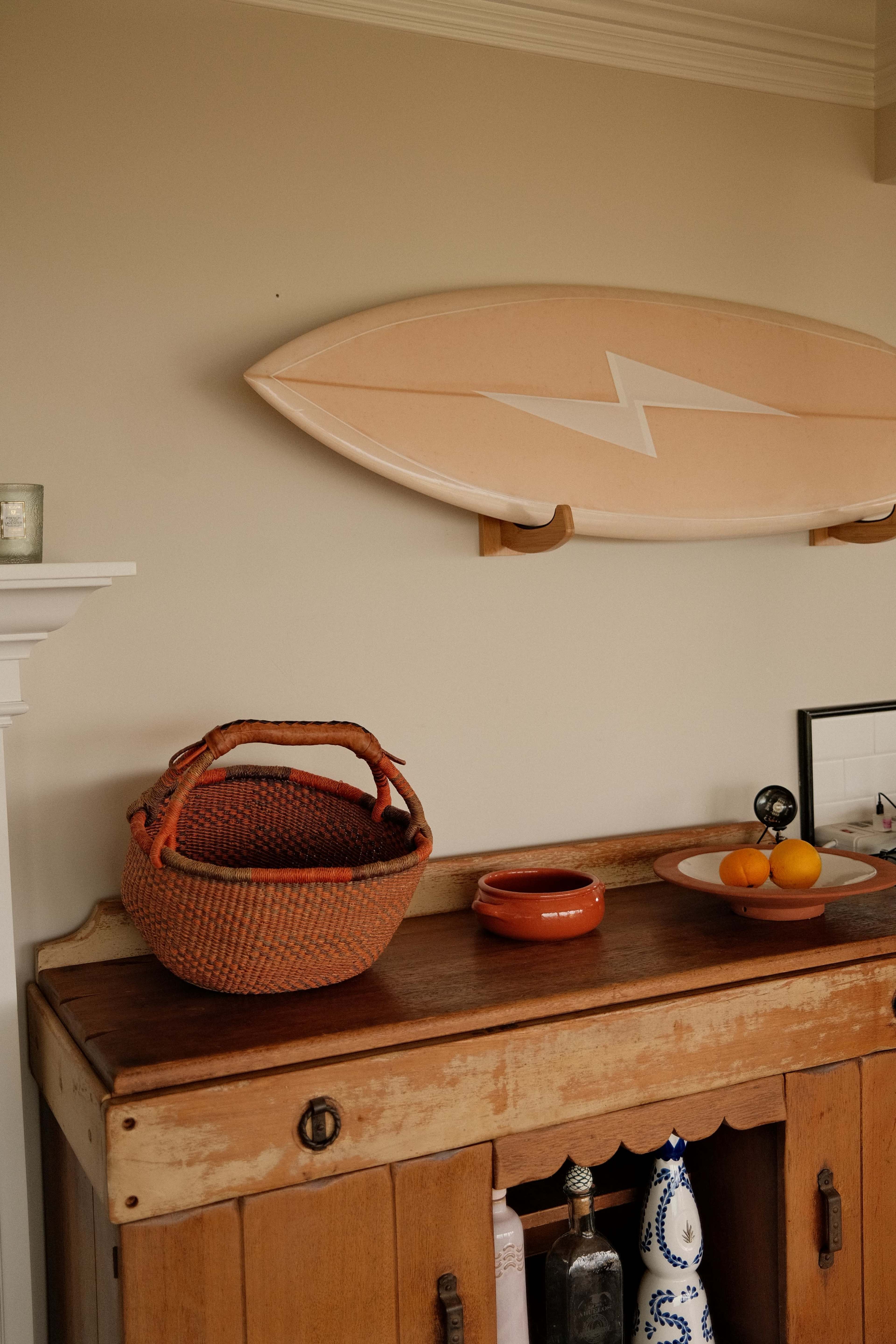 A wooden sideboard displays a woven basket, a small bowl, and fresh oranges beneath a surfboard mounted on the wall.