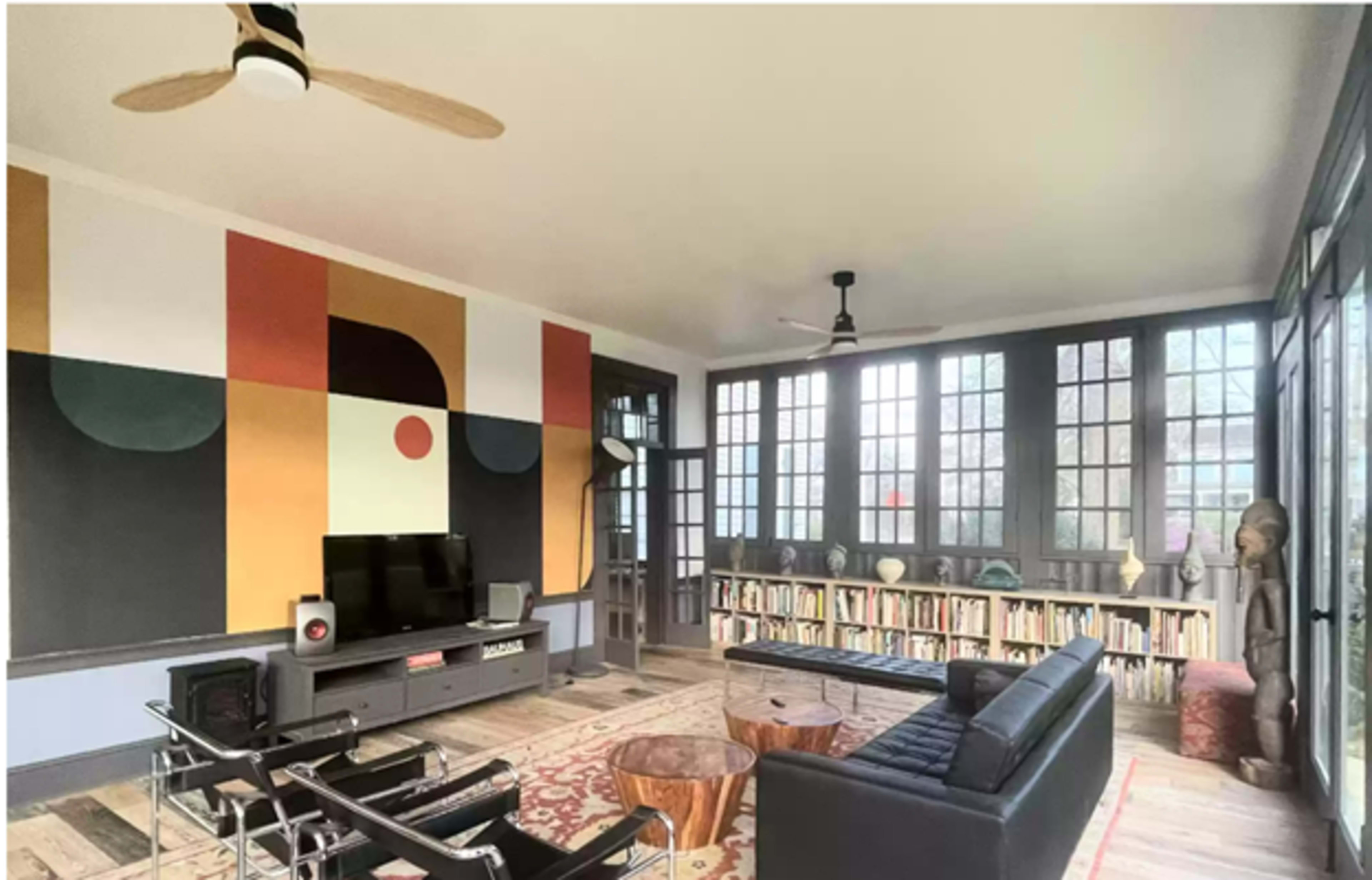 A modern living room with large windows, featuring a black sofa, a wooden coffee table, a TV on an entertainment console, and a bookshelf against the wall, all set against a colorful abstract mural.