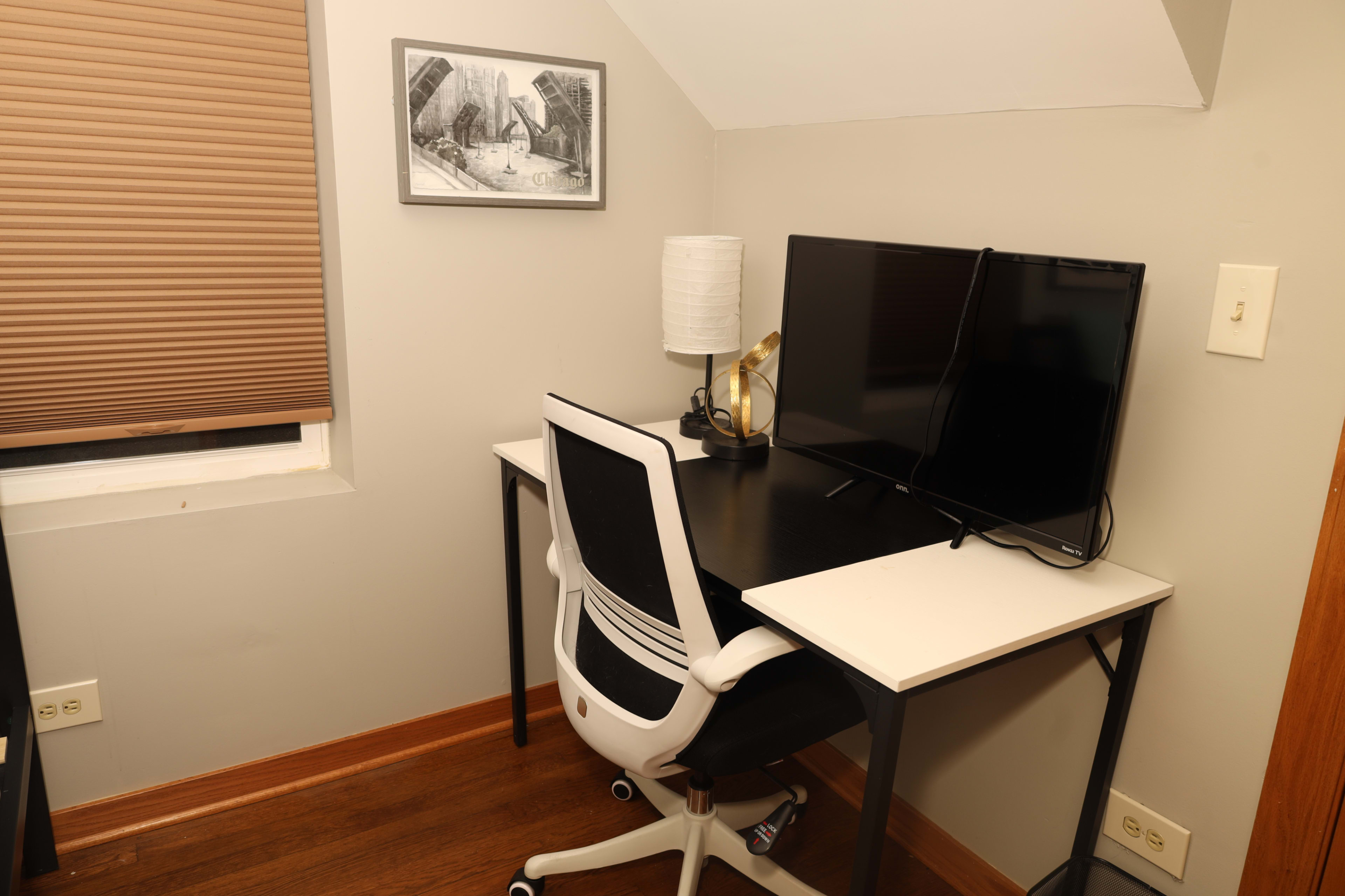 The image shows a modern workspace featuring a desk with a monitor, a white office chair, a lamp, and a black-and-white framed artwork on the wall.