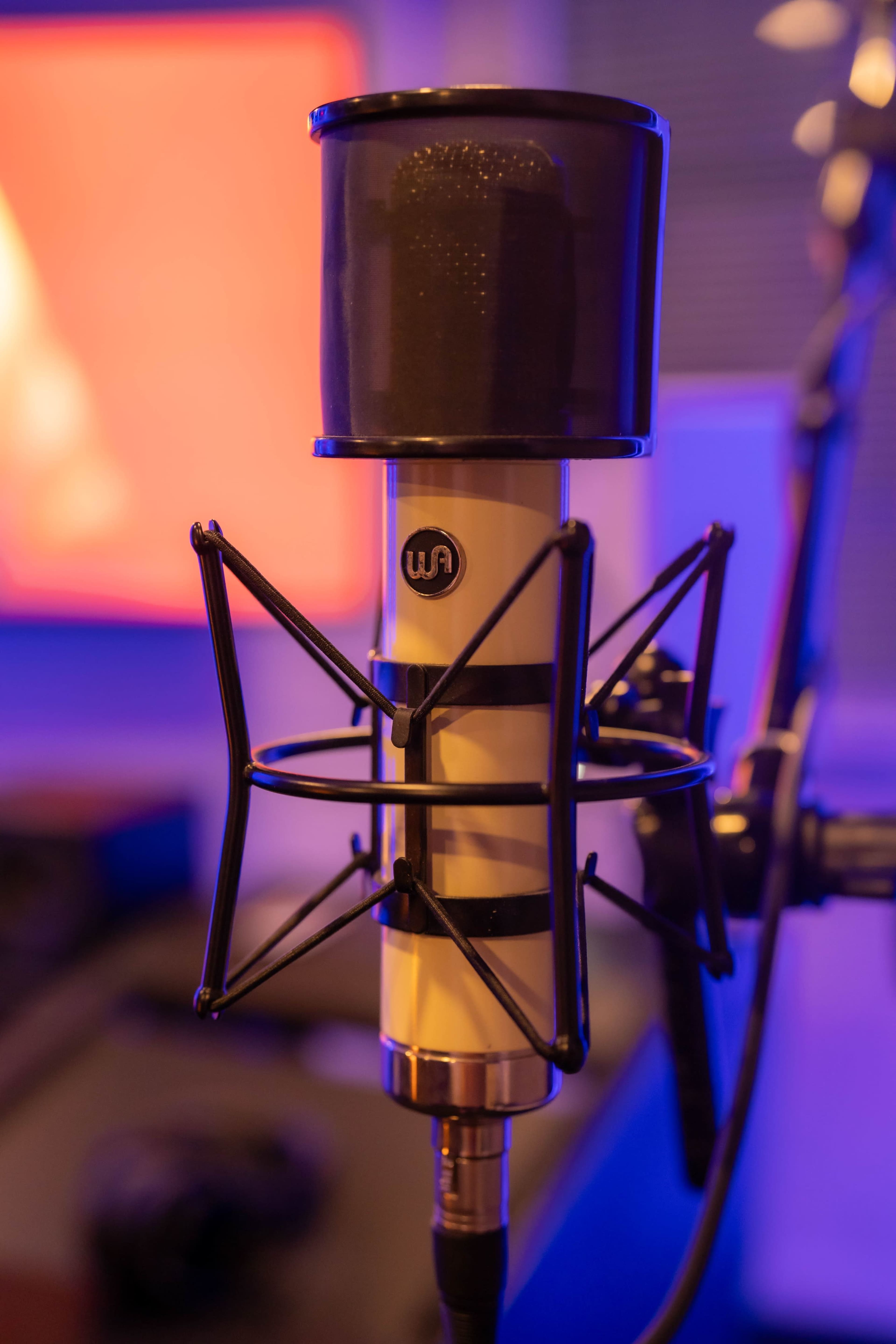 A close-up of a microphone with a black mesh pop filter and a shock mount, set against a blurred colorful background.