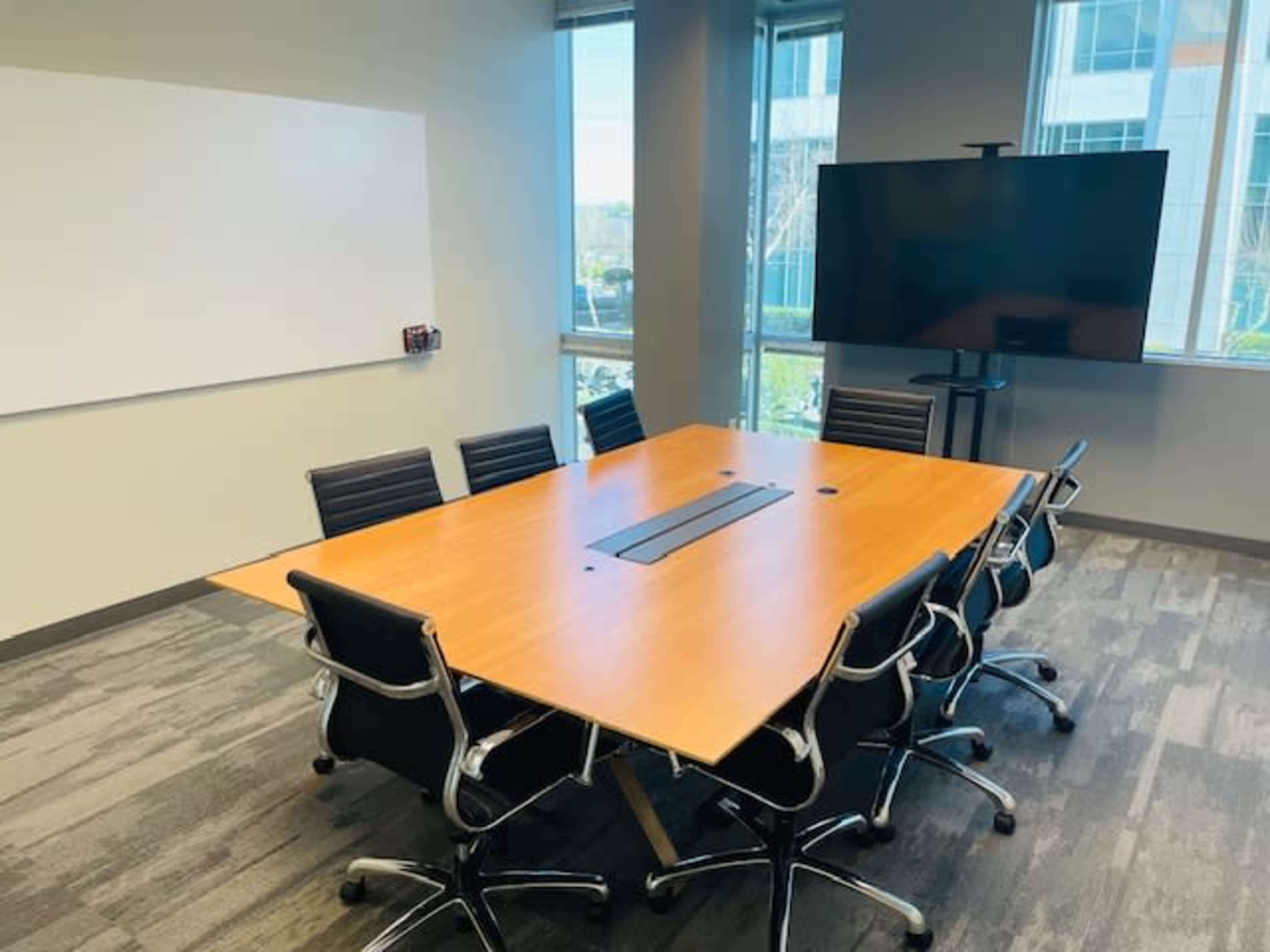 A conference room features a large wooden table surrounded by eight black chairs, with windows providing natural light and a large screen on one wall.