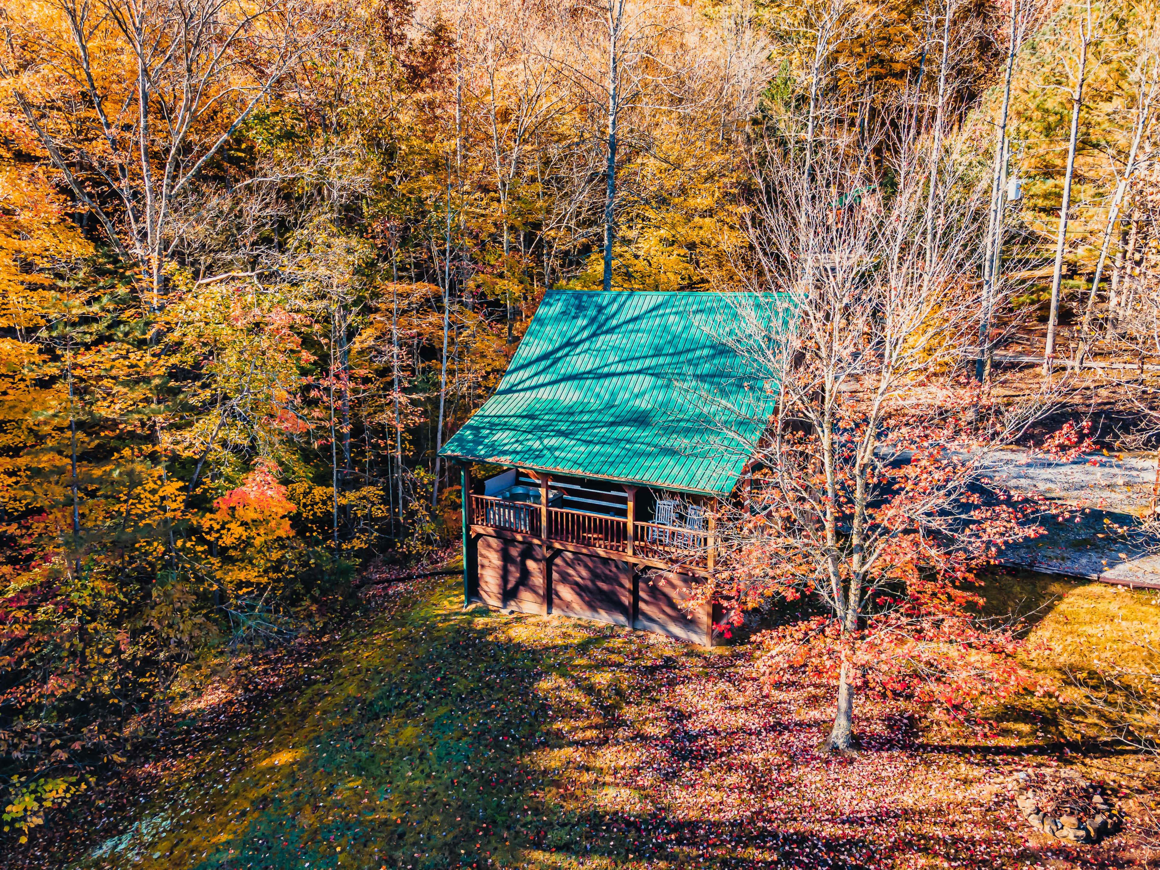 A wooden cabin with a green roof is surrounded by colorful autumn foliage in a forested area.