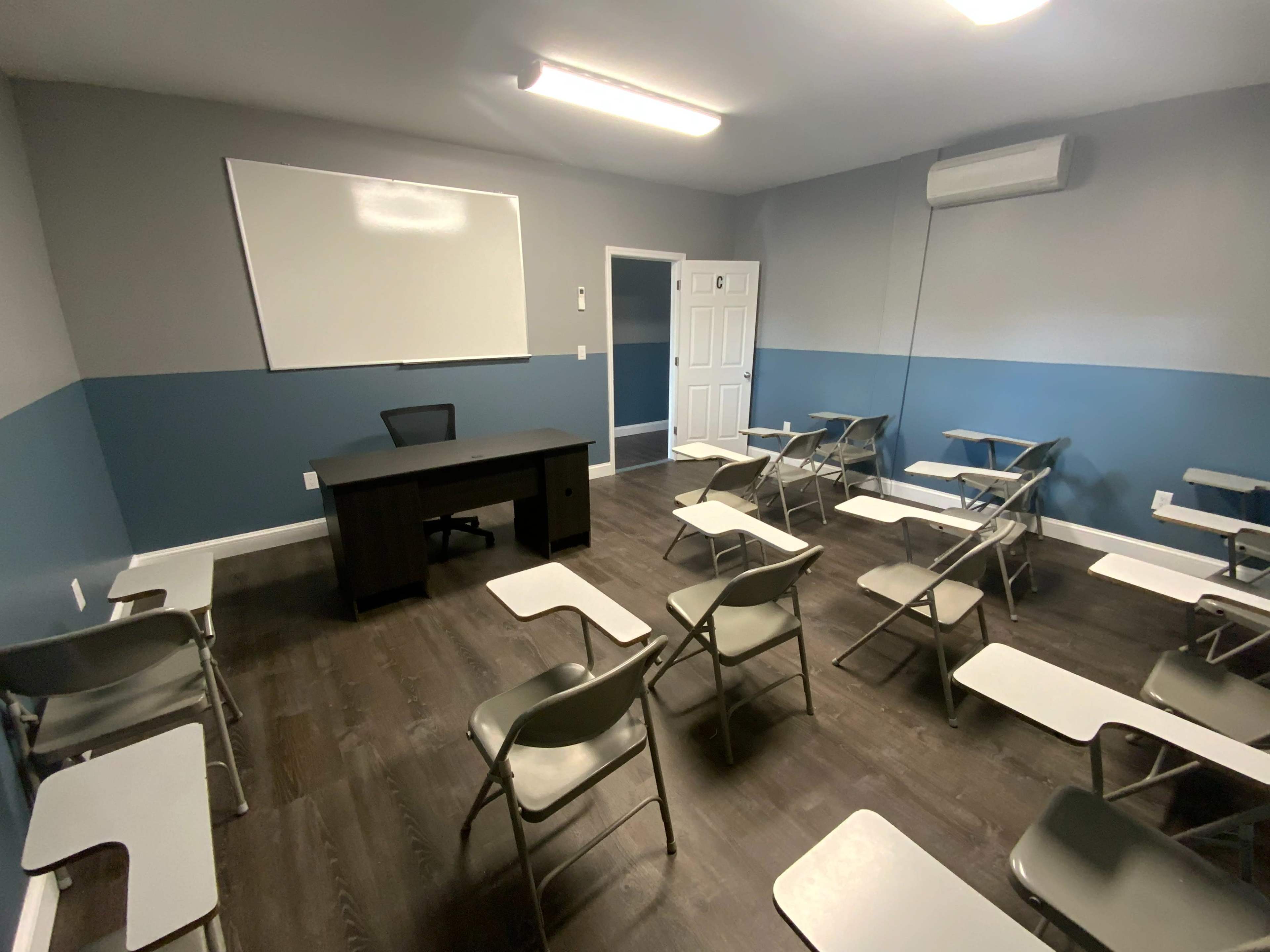 A classroom setting with several rows of folding chairs, a desk at the front, and a whiteboard on the wall.