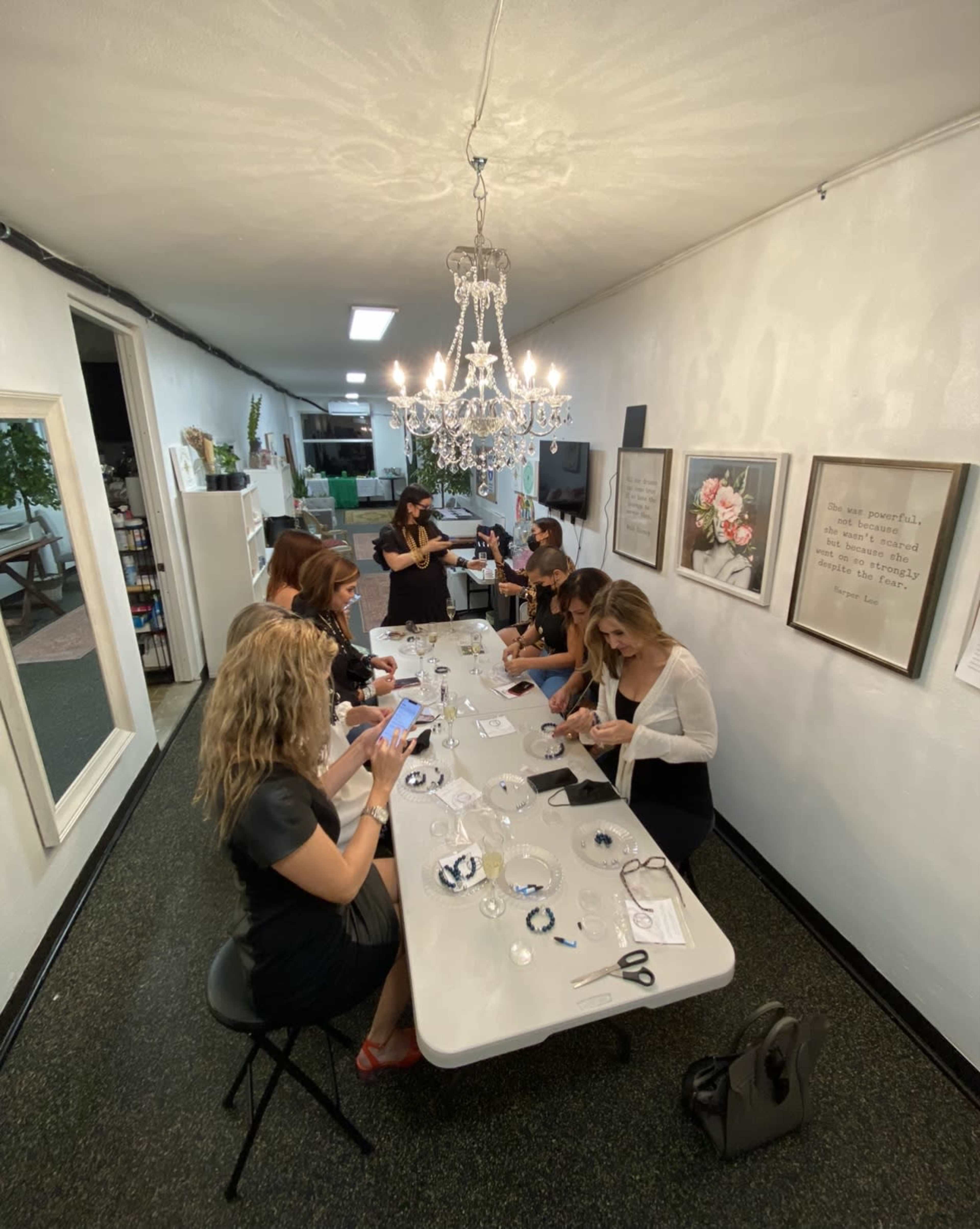 A group of women is engaged in a craft activity around a long table in a well-lit room with white walls and a chandelier overhead.