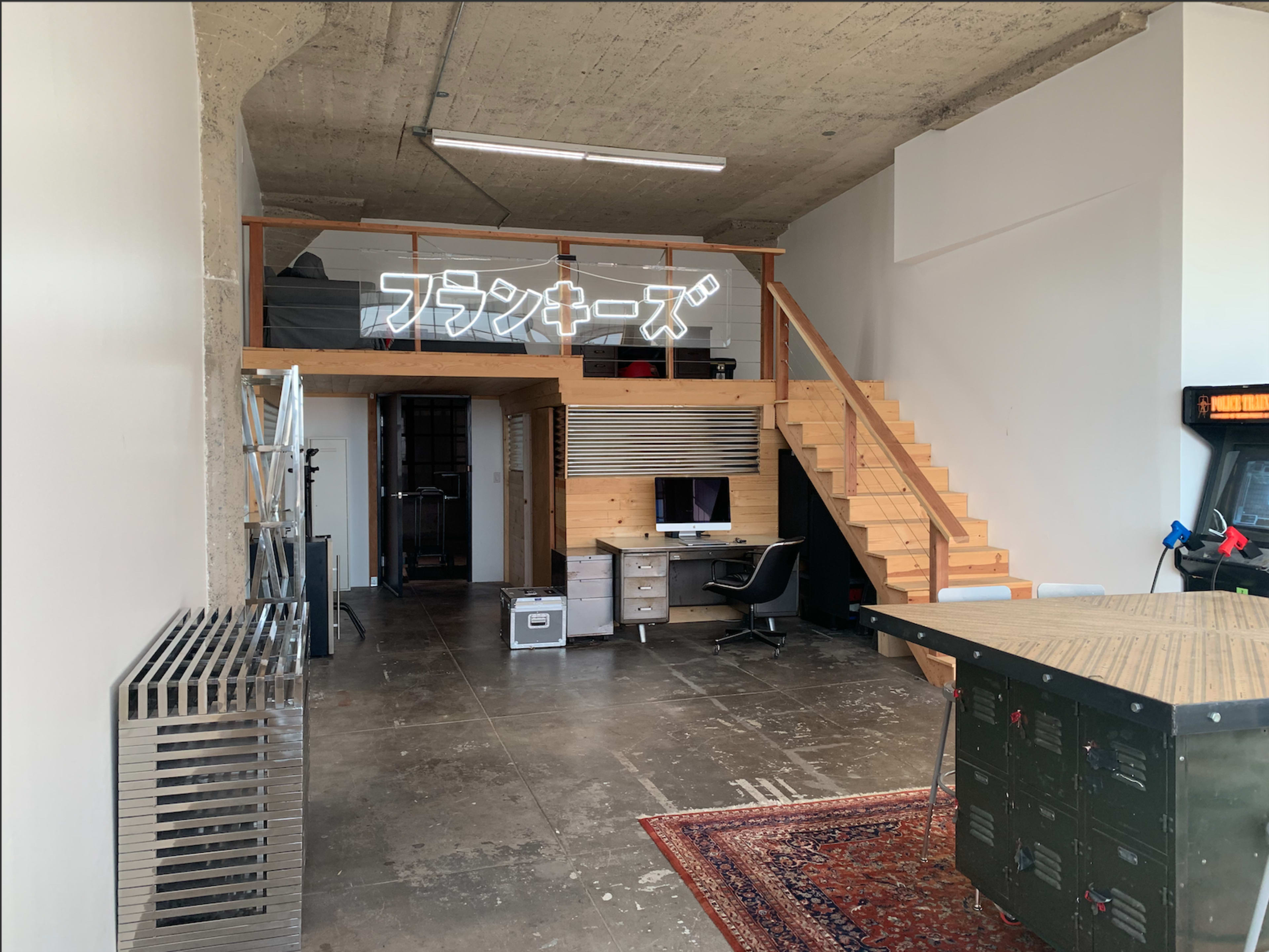 A modern, open-plan office space featuring a wooden staircase, a desk with a computer, and a decorative sign in Japanese on the wall.