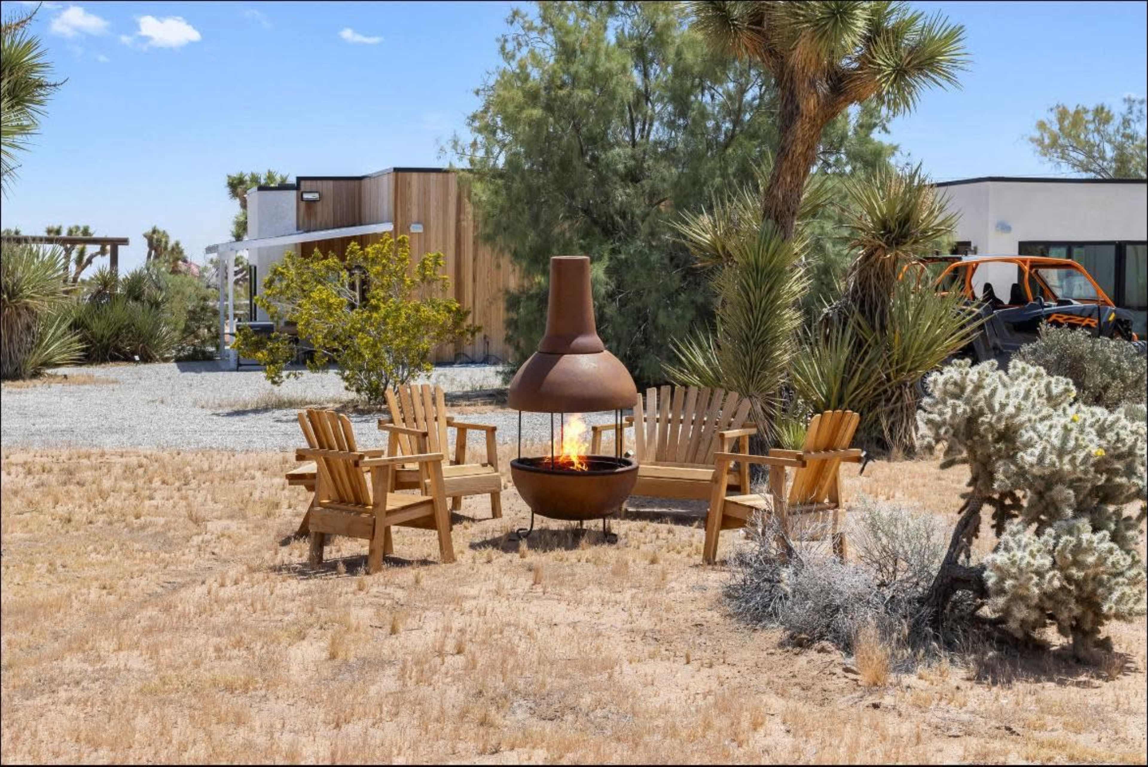 A circular seating arrangement of wooden chairs surrounds a metal fire pit in a desert landscape near a modern house.
