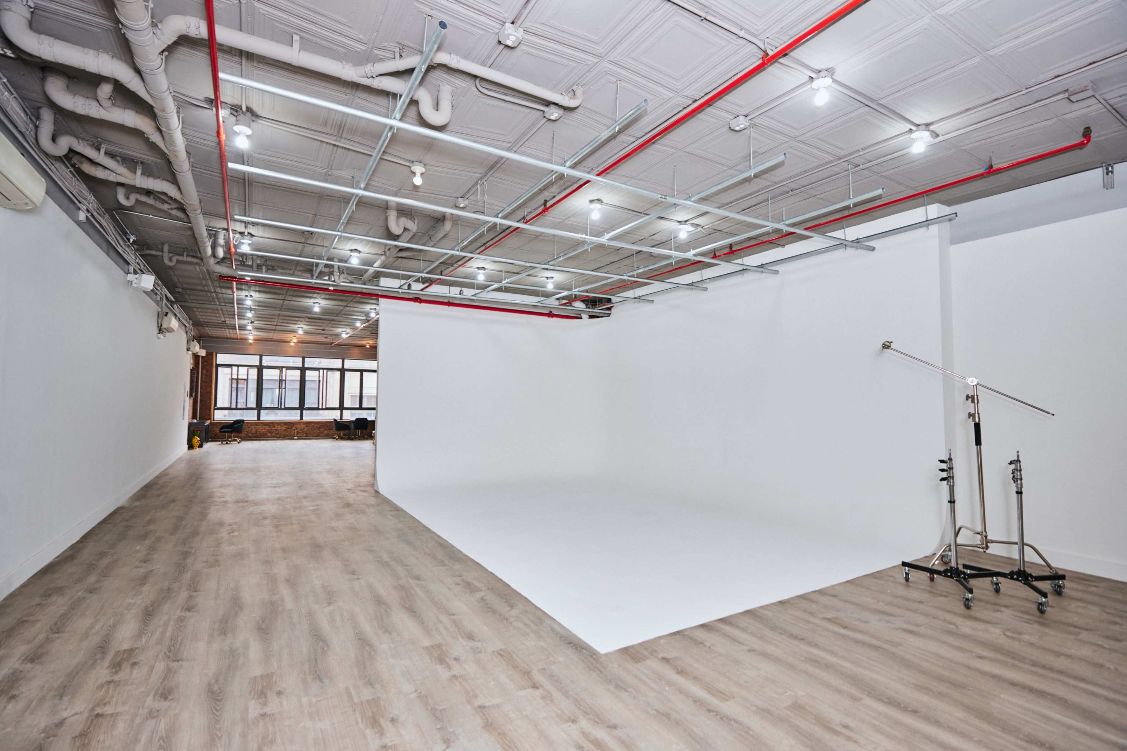 A spacious studio with a white backdrop, wooden flooring, and overhead lighting along with exposed pipes and a rolling stand.
