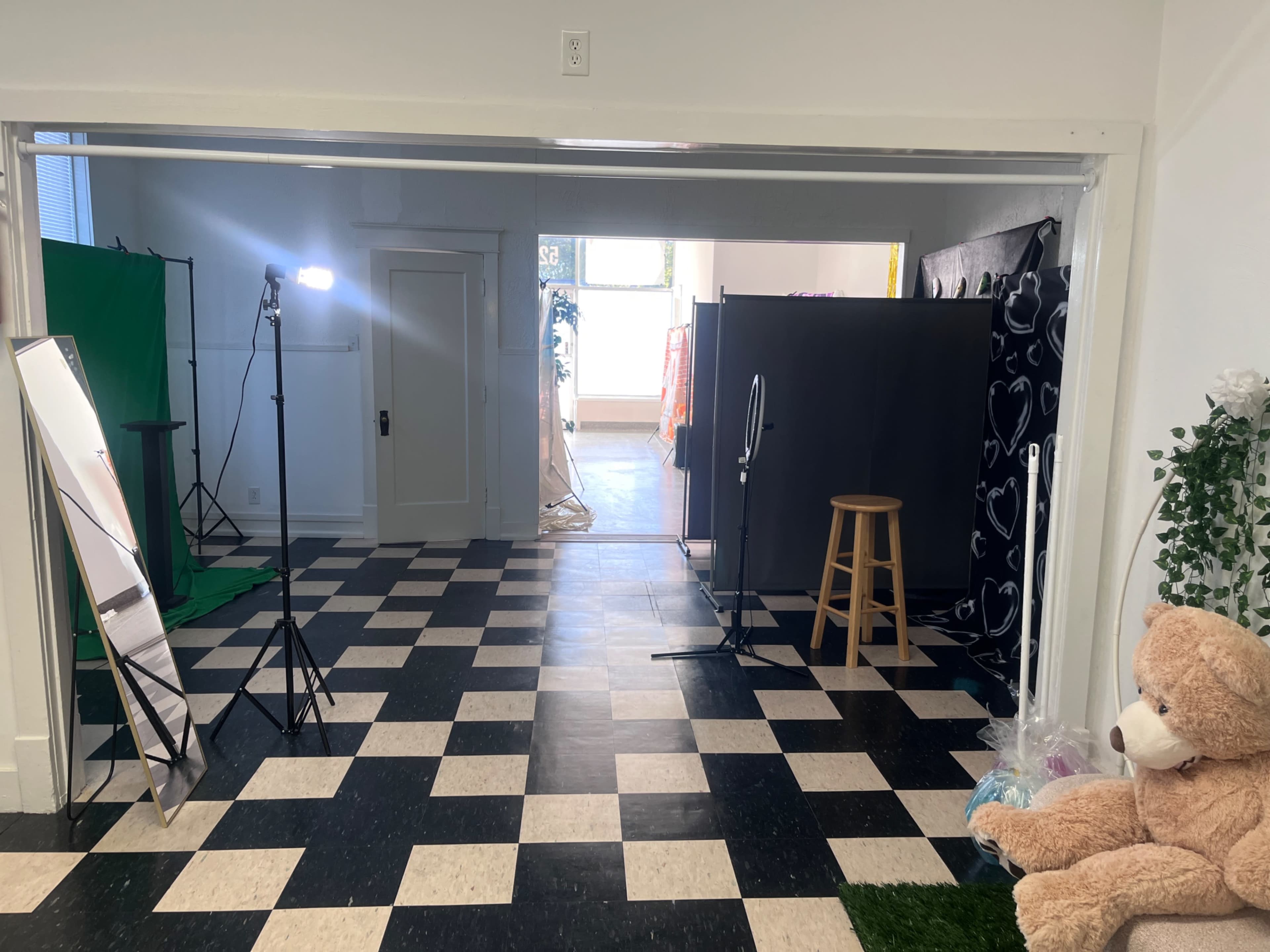 The image shows a studio space divided by a black curtain, with a light setup, a mirror, a stool, and a large teddy bear in the foreground.