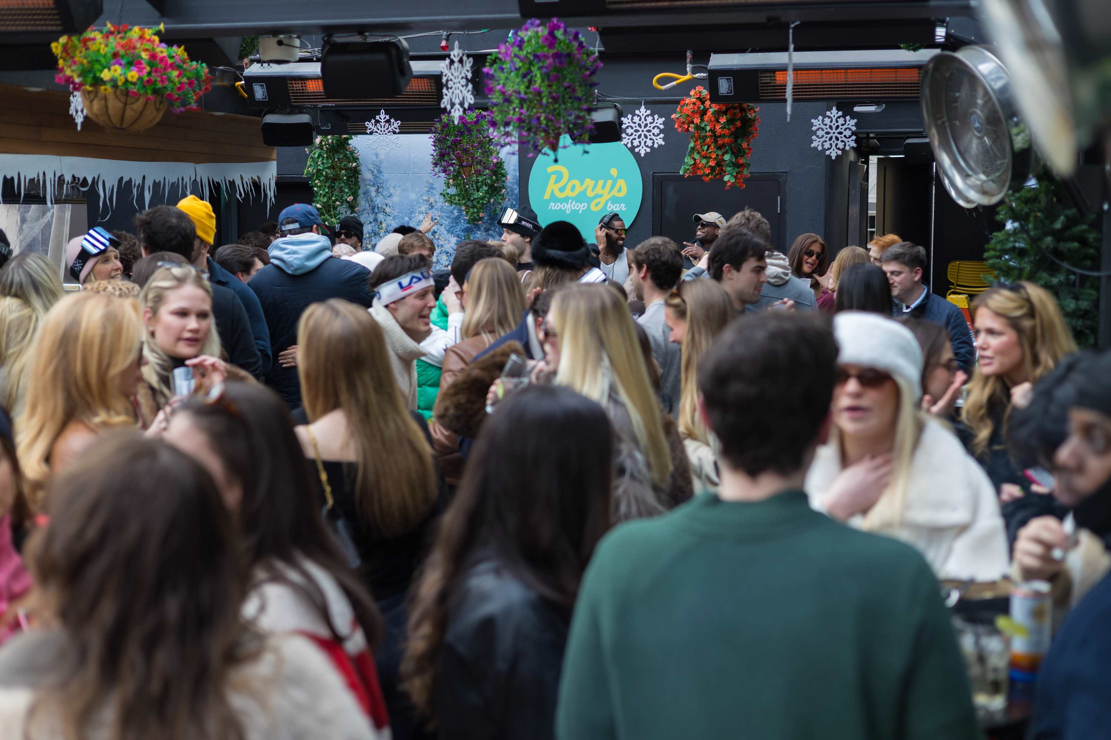 A large crowd of people mingles at a lively outdoor event, surrounded by decorations that include colorful flowers and snowflakes.