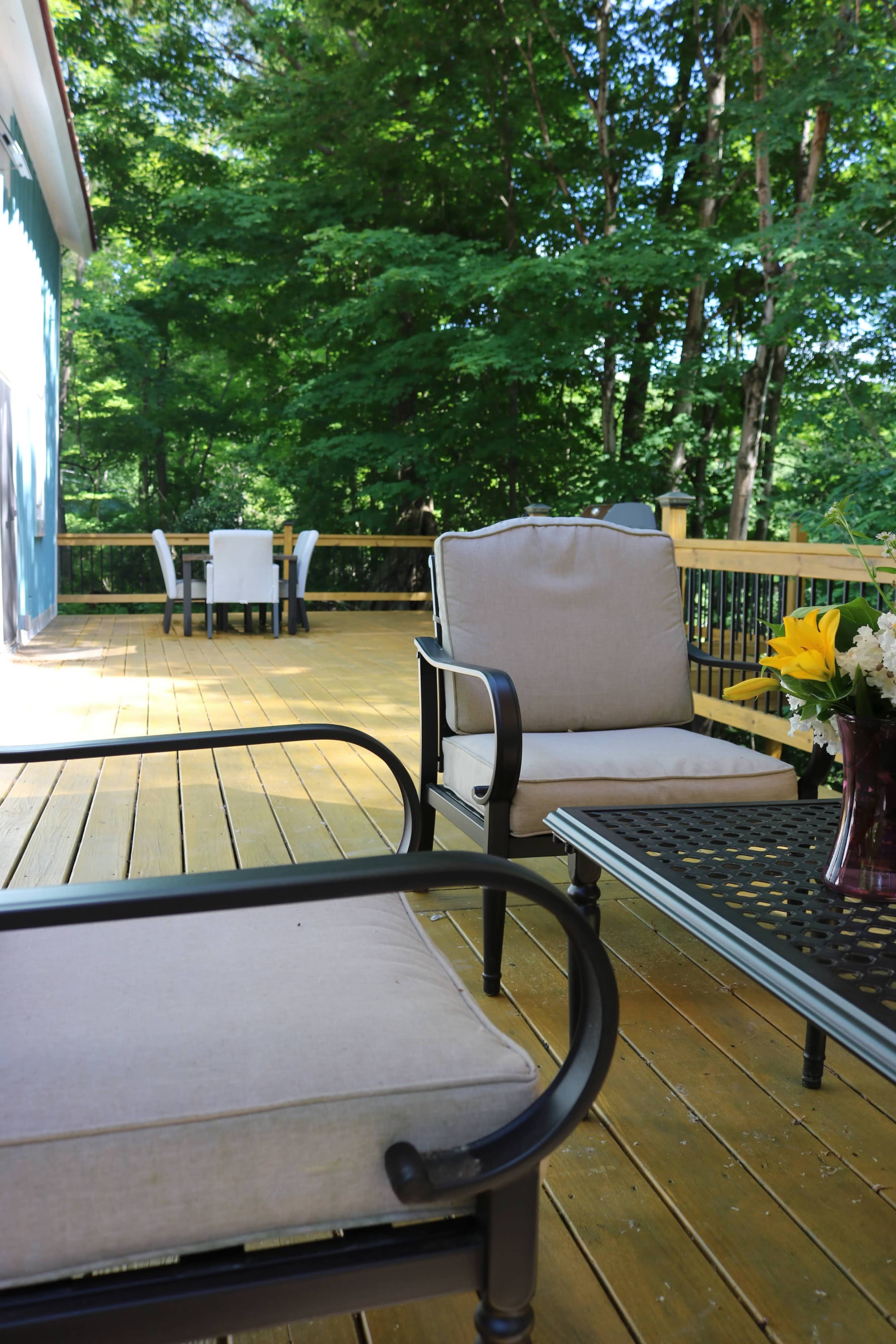 The scene shows a wooden deck with two lounge chairs, a coffee table, and a dining area set up with chairs in the background, surrounded by greenery.