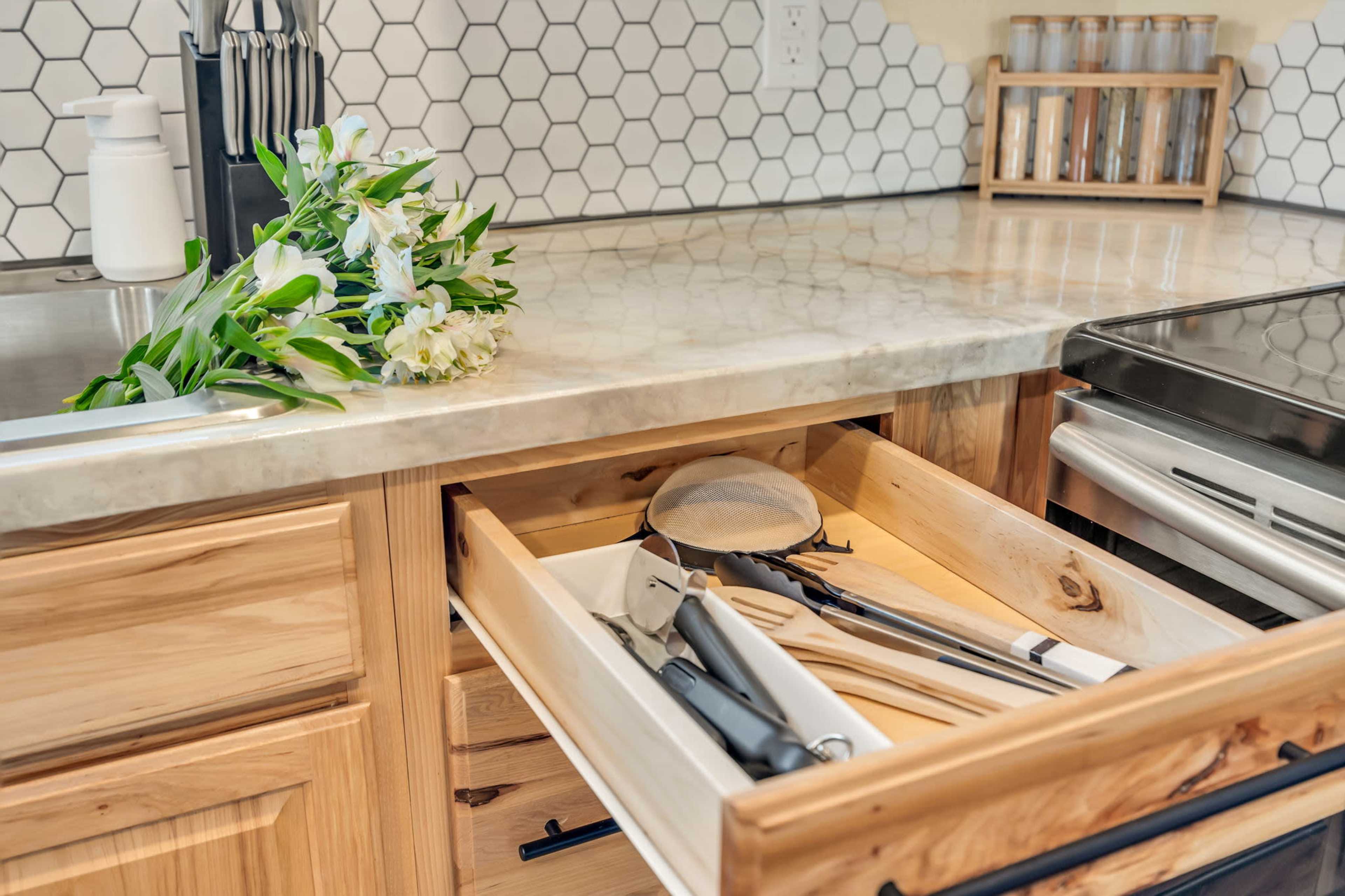 A wooden kitchen drawer is partially open, revealing cooking utensils and a strainer, while fresh flowers rest on the countertop beside a hexagonal tile backsplash.