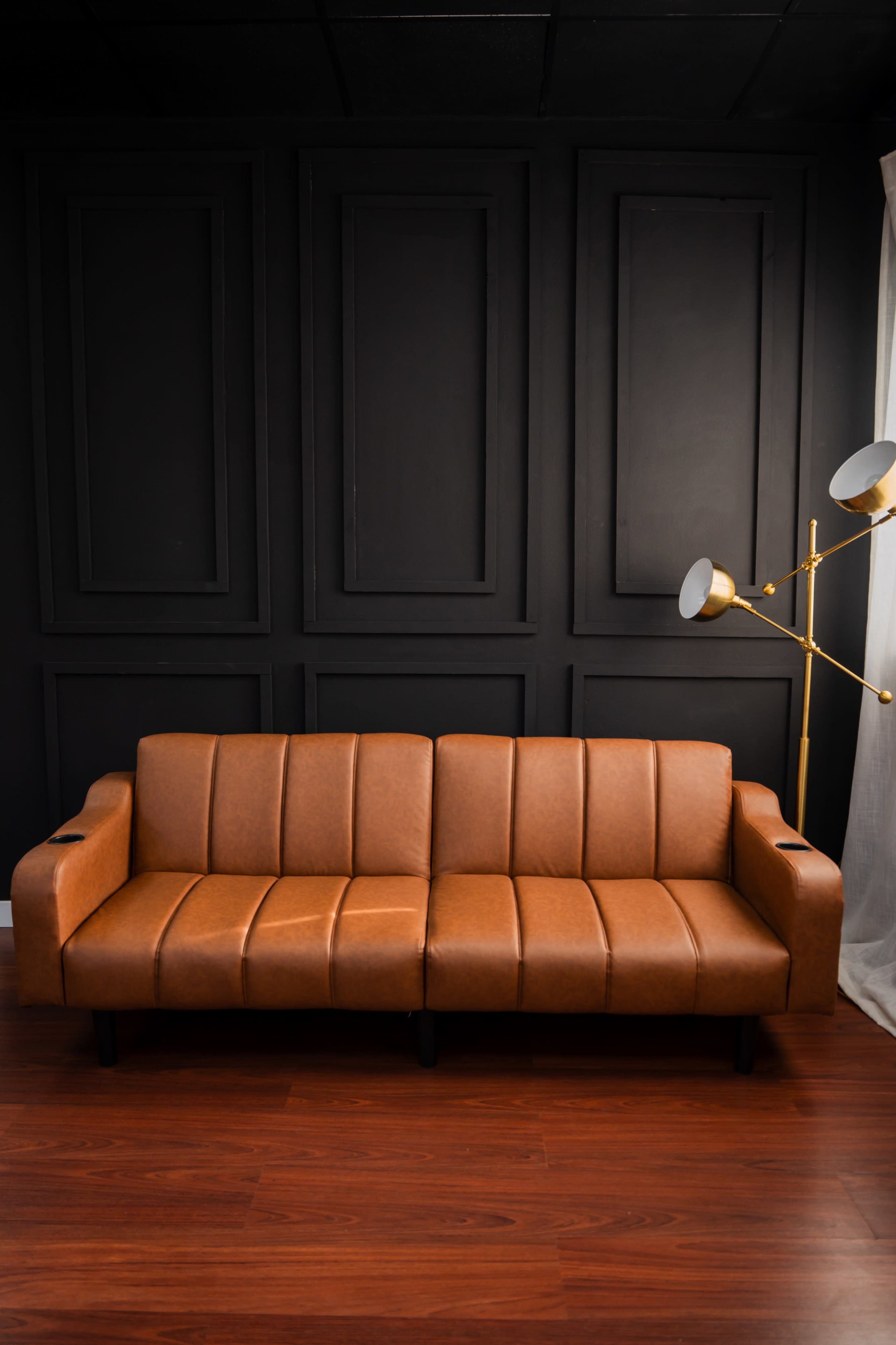 The image features a brown leather sofa with vertical stripes, positioned against a black-paneled wall and accompanied by a floor lamp.