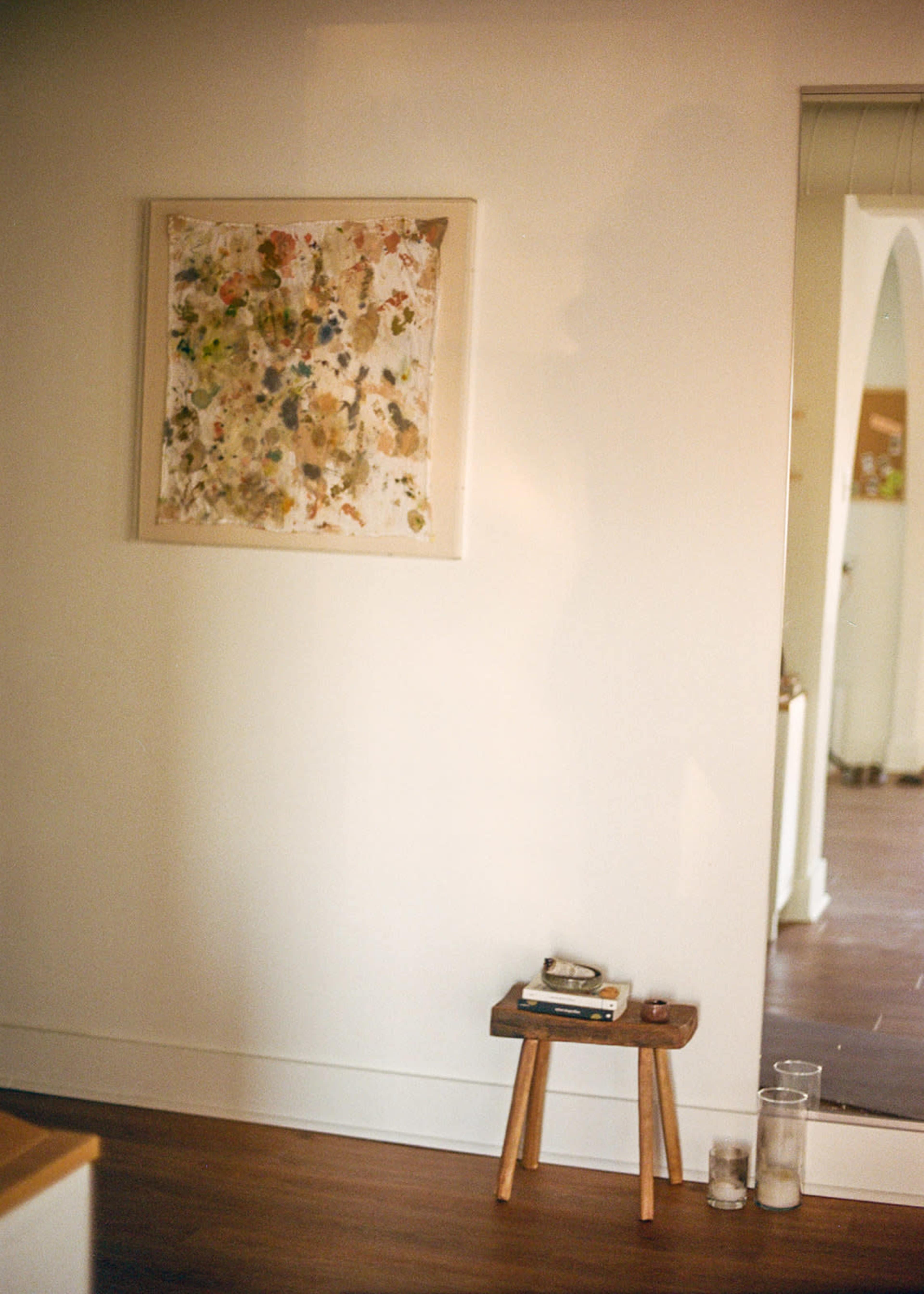 A framed abstract painting hangs on a white wall beside a small wooden stool topped with books and two glass jars.
