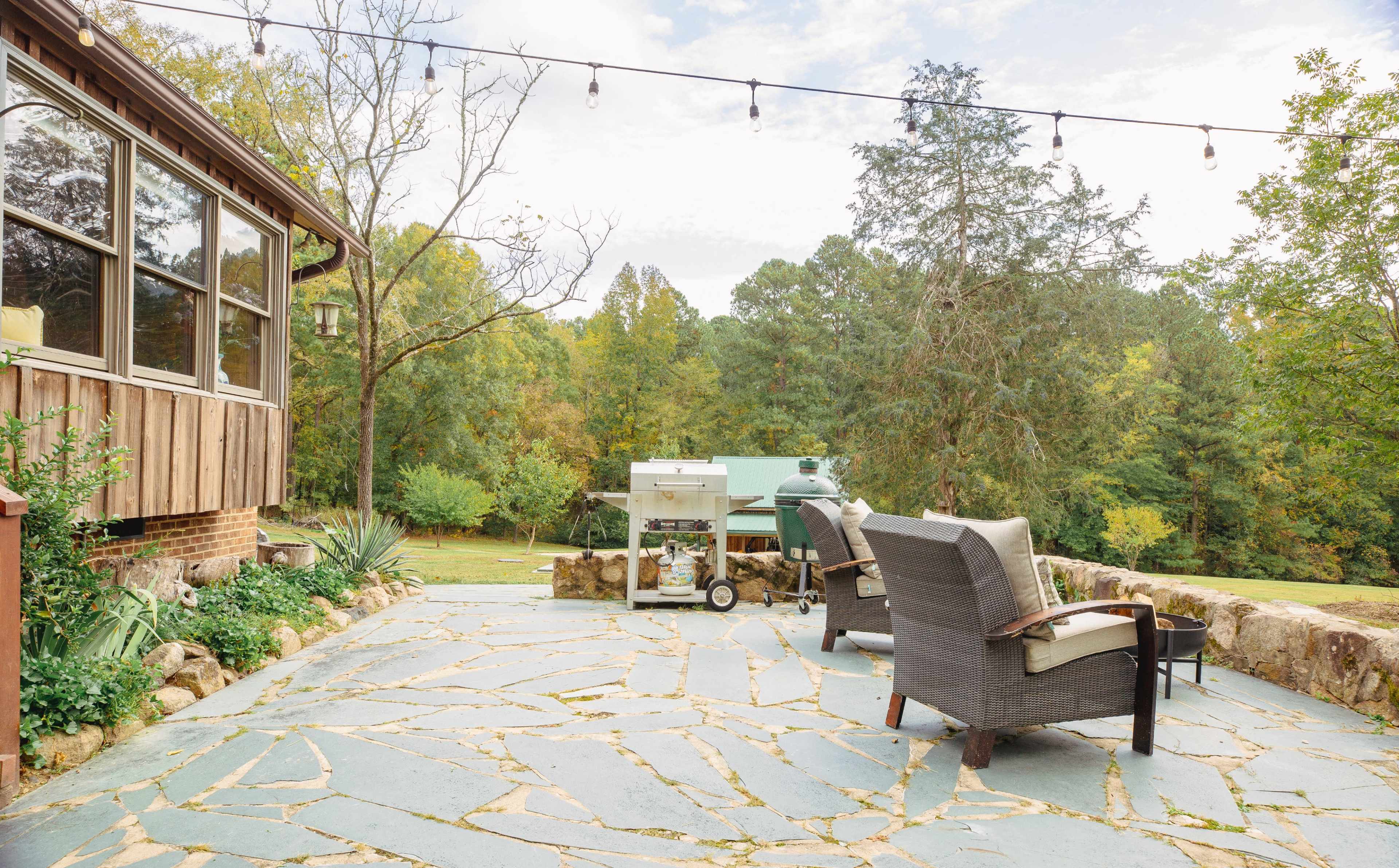 A stone patio features two chairs and a grill, surrounded by greenery and trees.