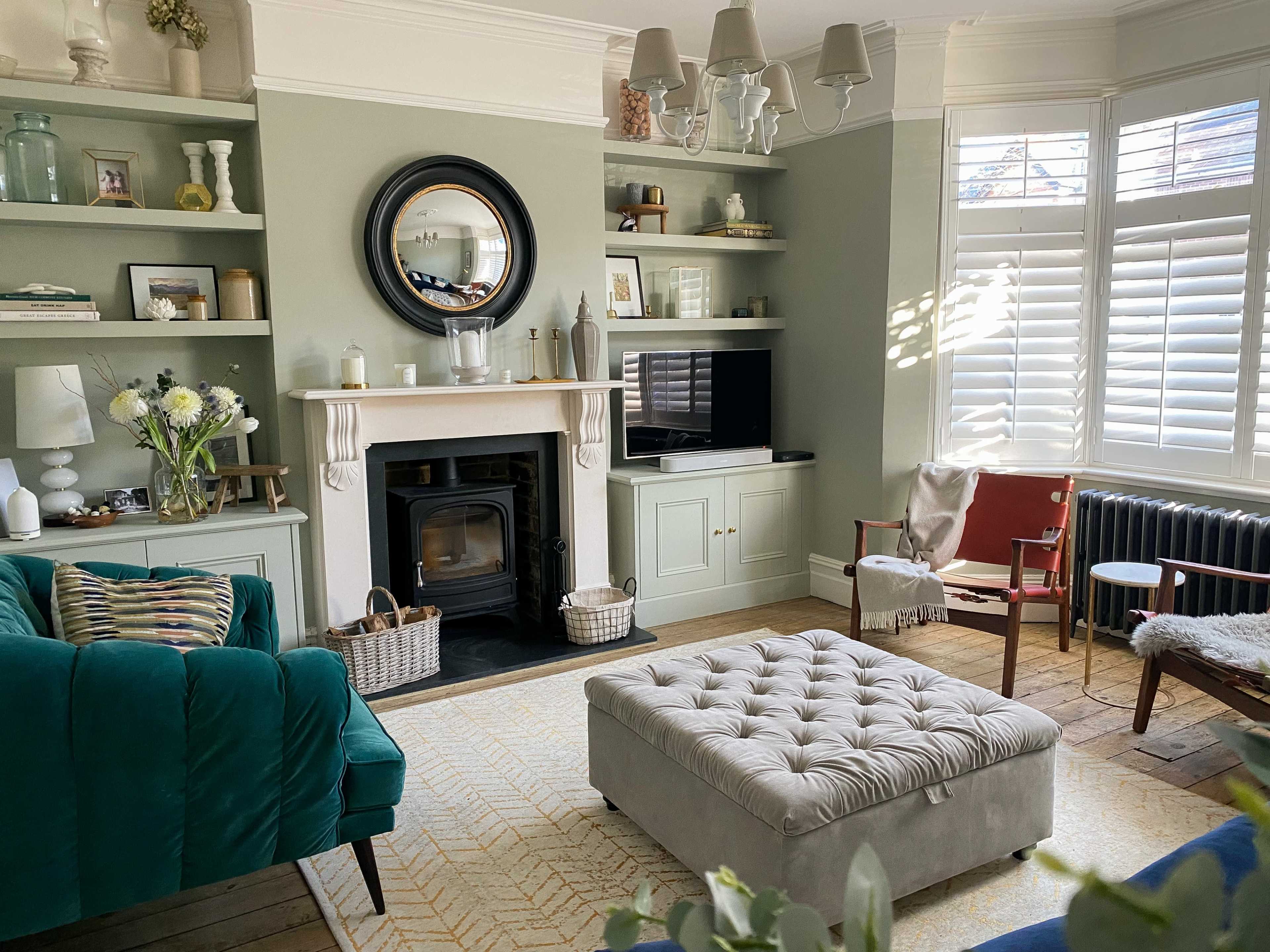 A stylish living room with a green accent wall, built-in shelves filled with decor, a fireplace with a mirror above it, and a tufted gray ottoman on a patterned rug.