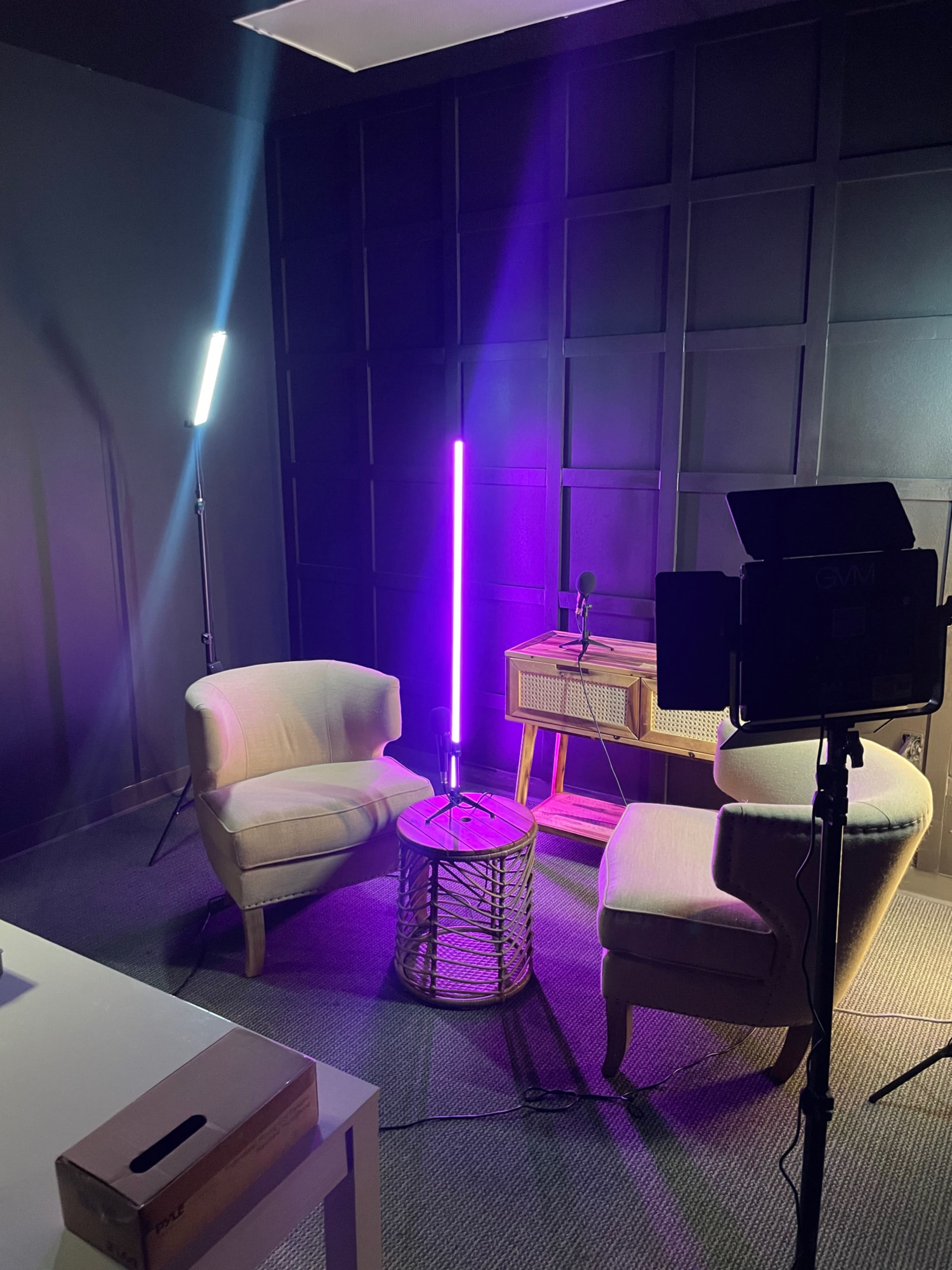 A small studio setup with two light sources, two chairs, a table made of woven material, and a microphone on a stand, all against a dark wall.