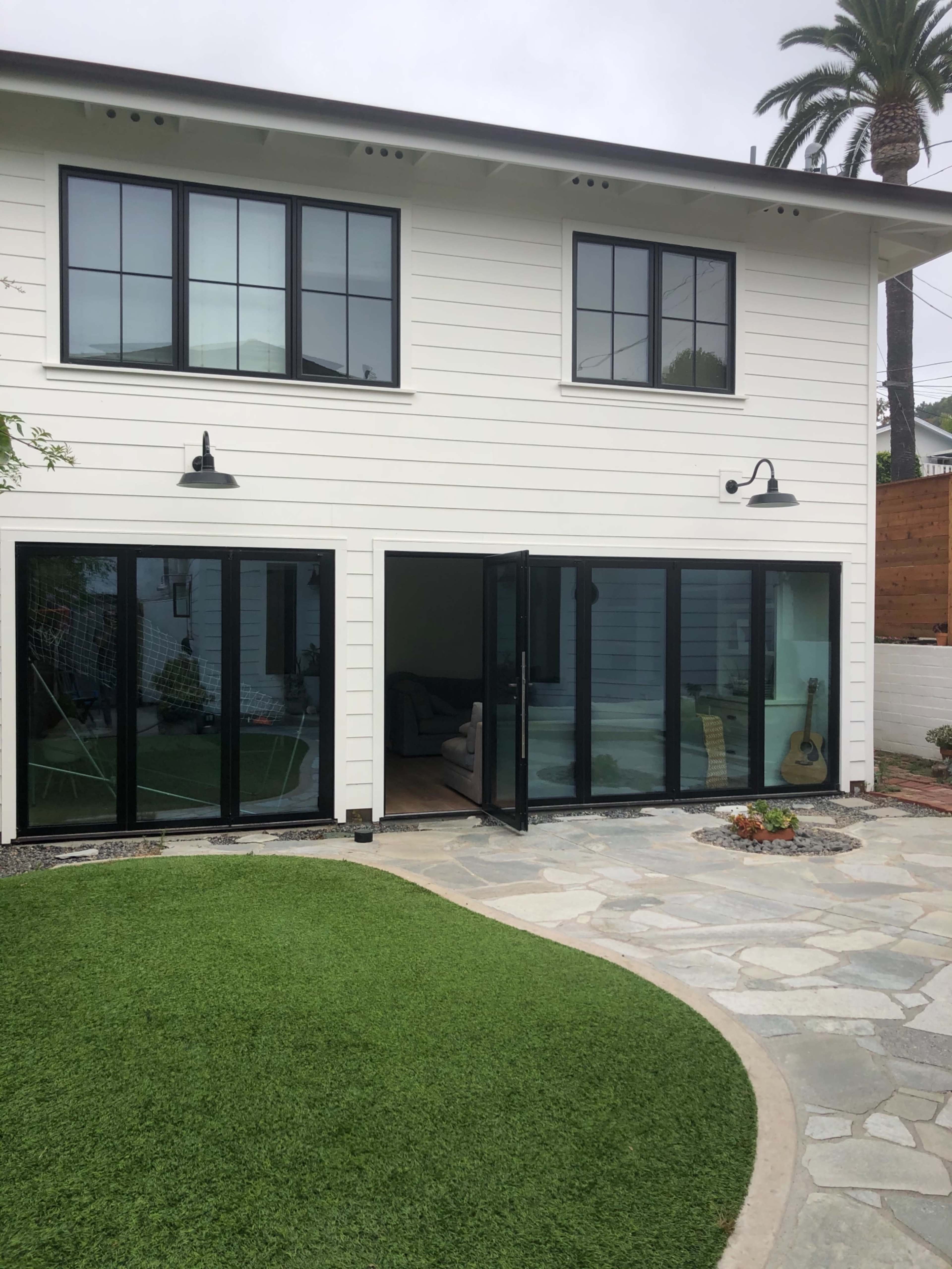A modern two-story house features large glass doors opening to a stone patio and a well-maintained lawn.