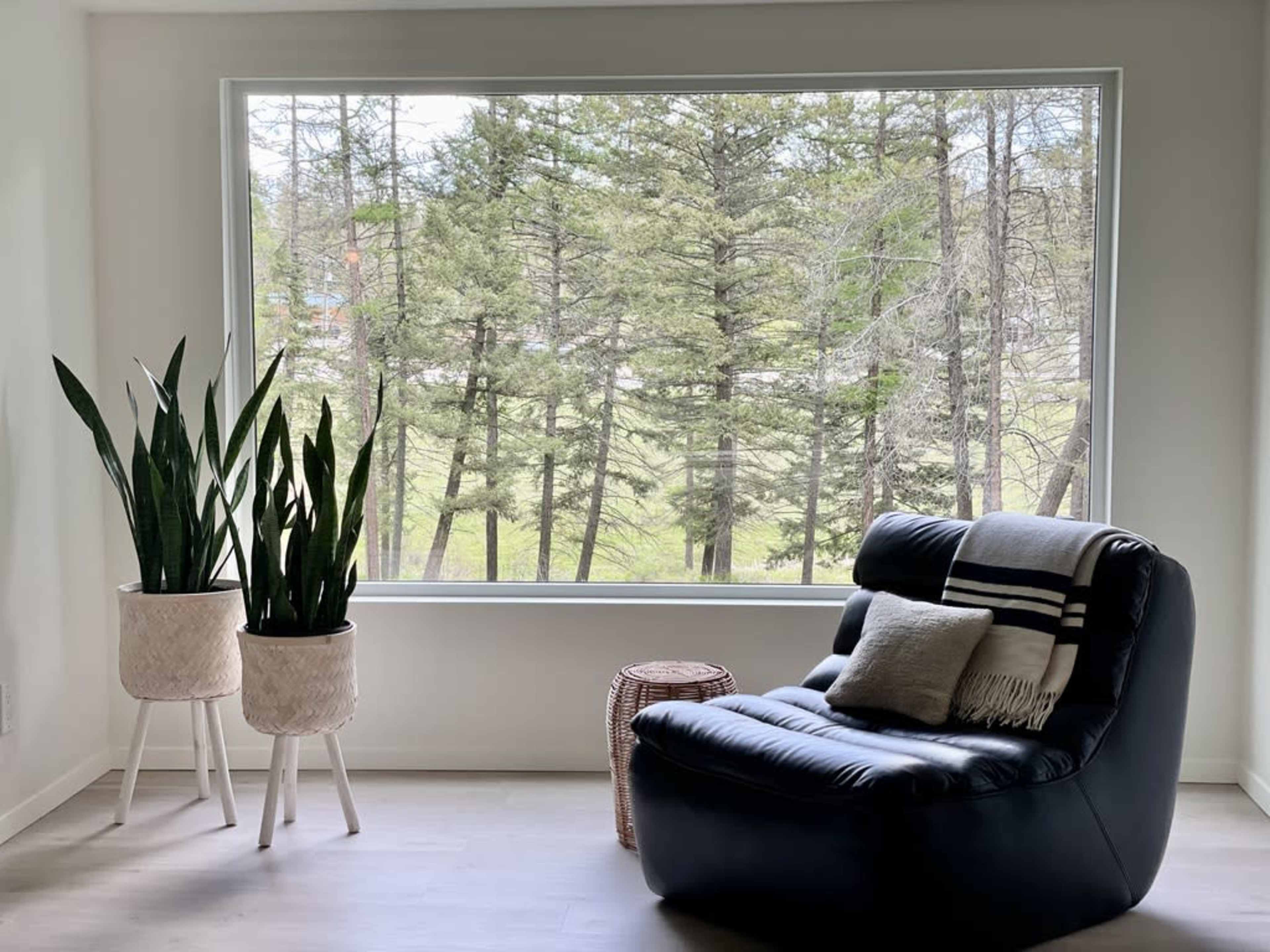 A black armchair with a cushion sits next to a round wicker table in front of a large window overlooking a grove of trees.