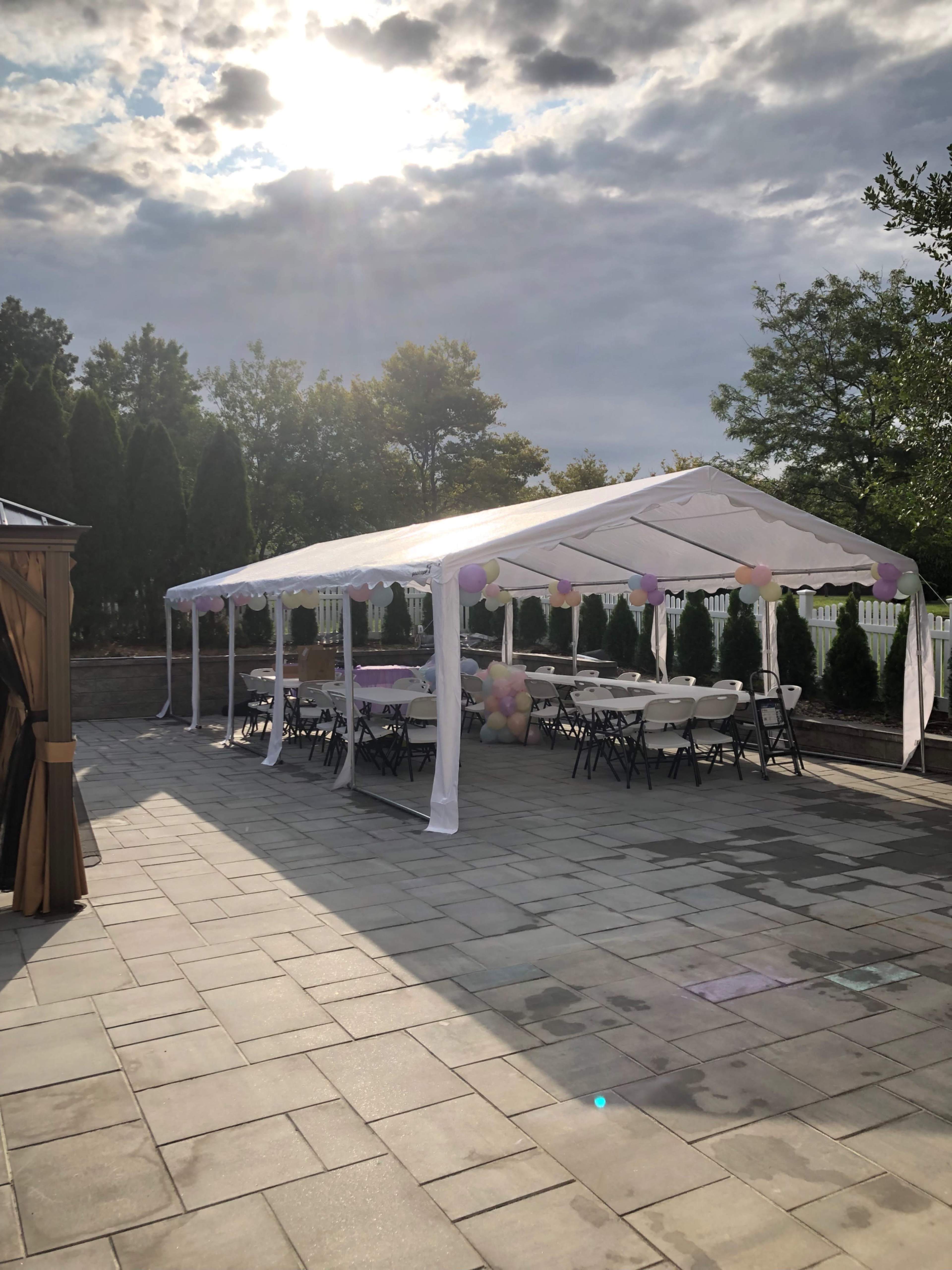 A white tent is set up on a stone patio, surrounded by greenery and decorated with balloons, ready for an event.