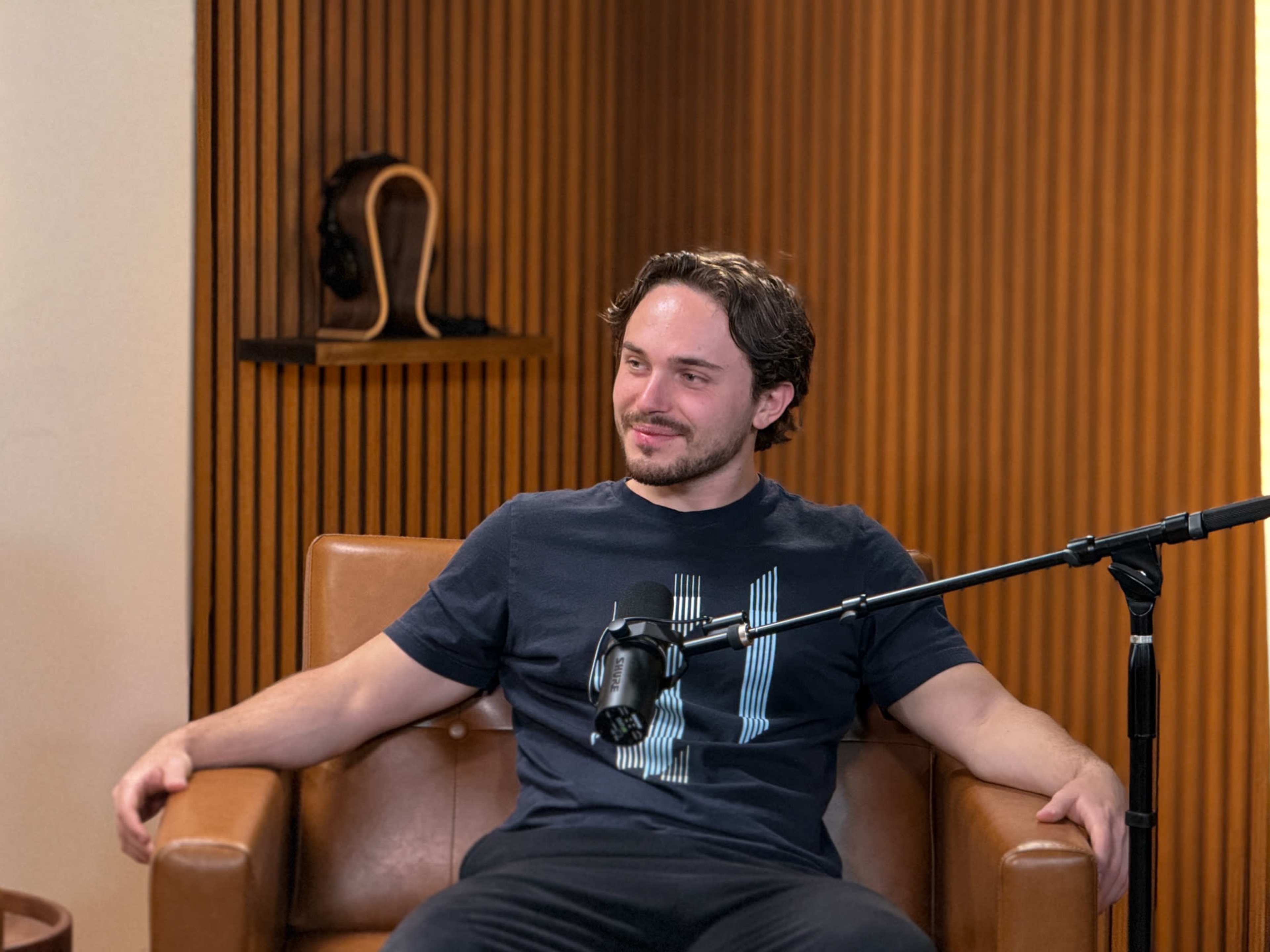 A man sits in a brown leather chair in a recording studio, smiling and holding a microphone attached to a stand.