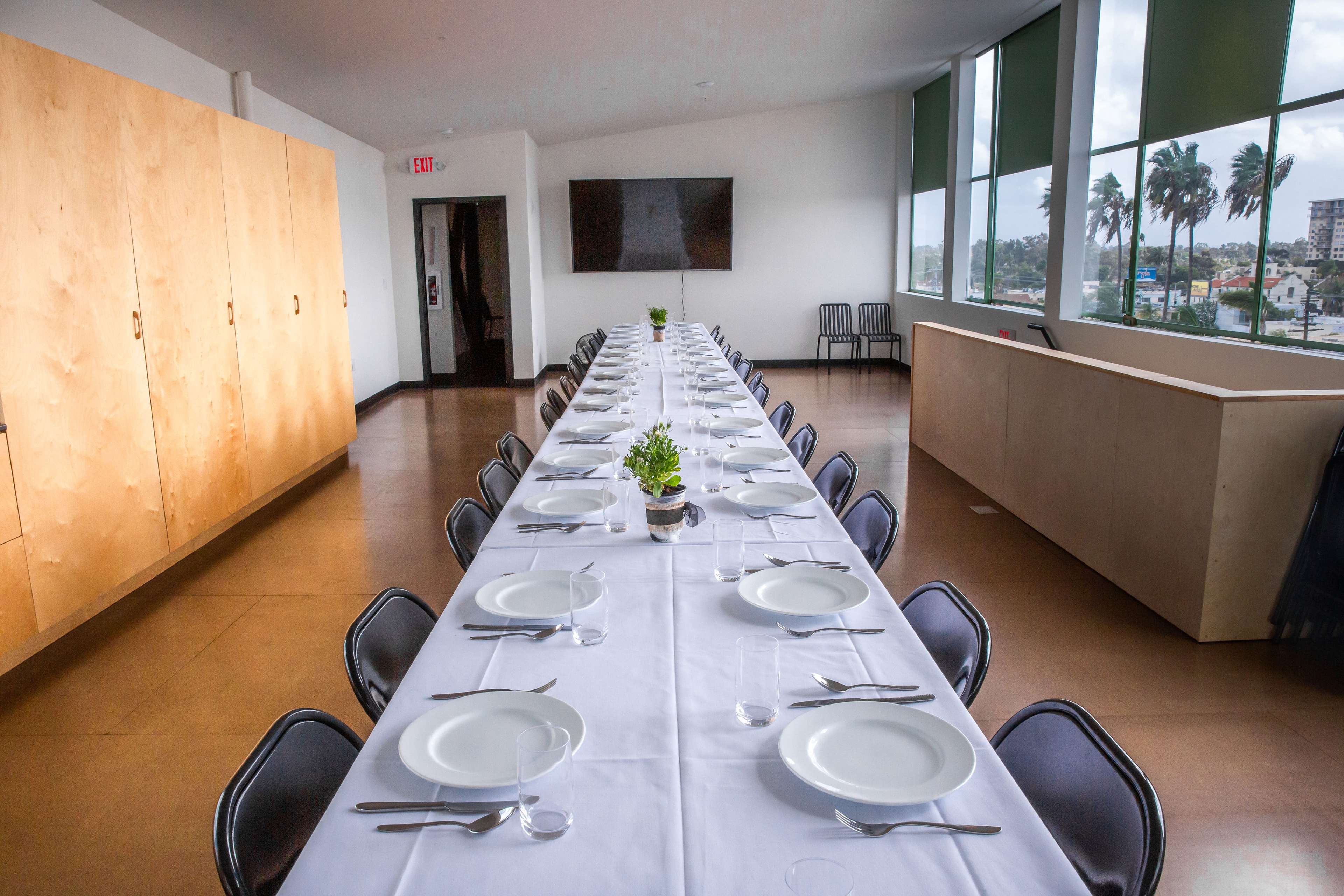 A long banquet table set with white plates and utensils is arranged in a brightly lit room with large windows and minimalist decor.