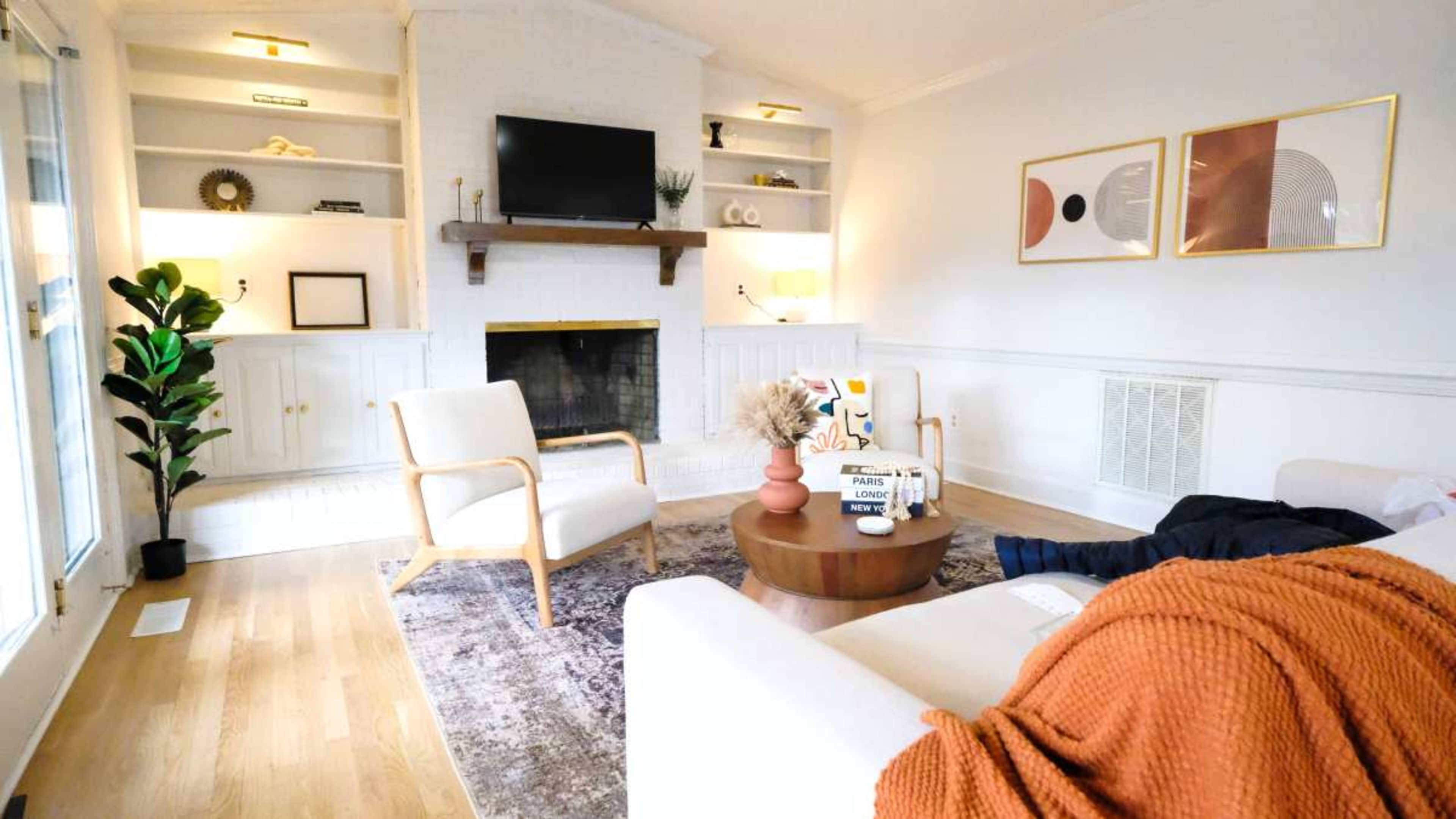 The image shows a modern living room with a fireplace, two beige armchairs, a round coffee table, and decorative plants.